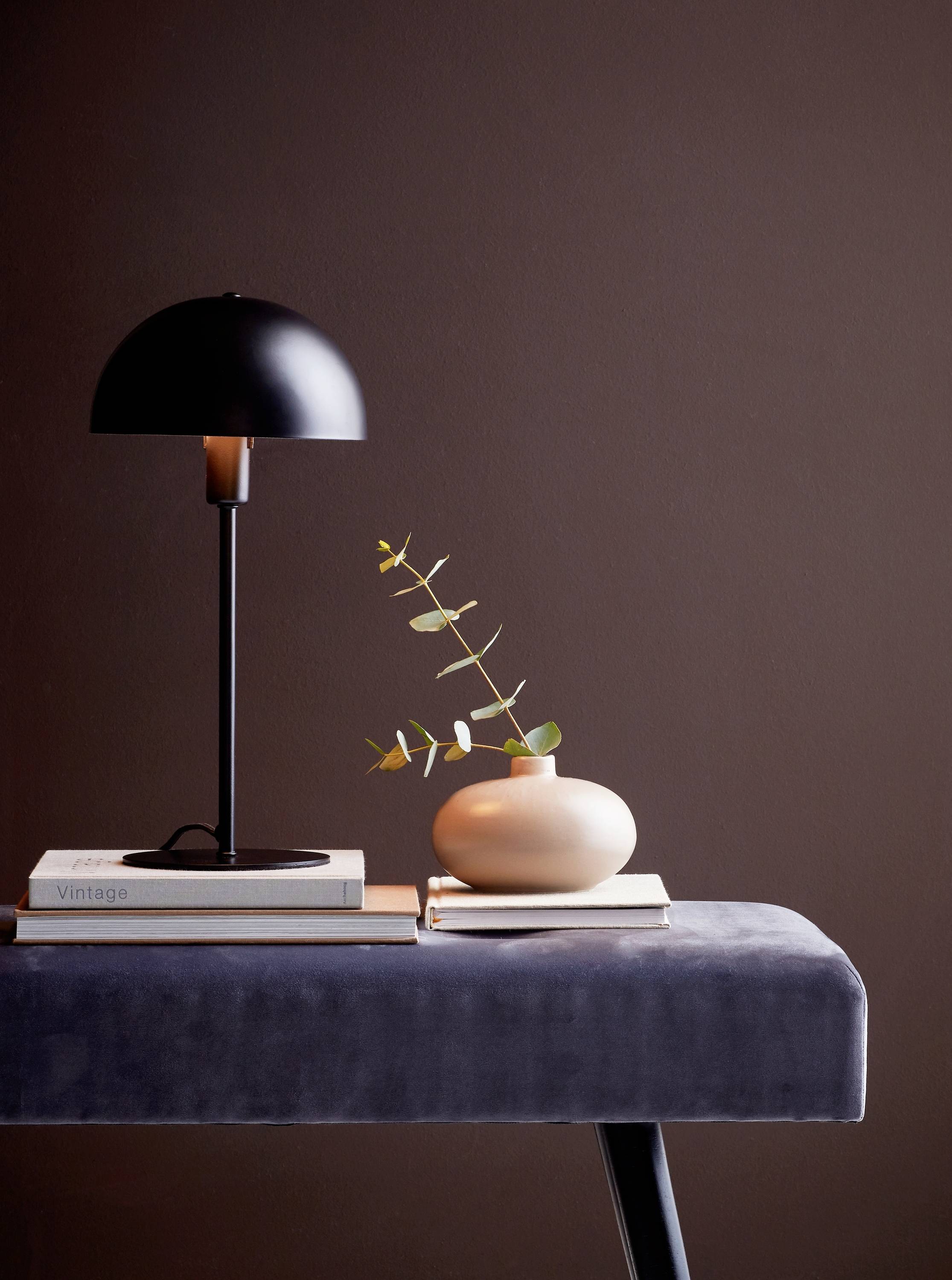 Stylowe martwe nature z czarną lampą, małym ceramicznym wazonem z gałązkami oraz kilkoma książkami na szarym stole.