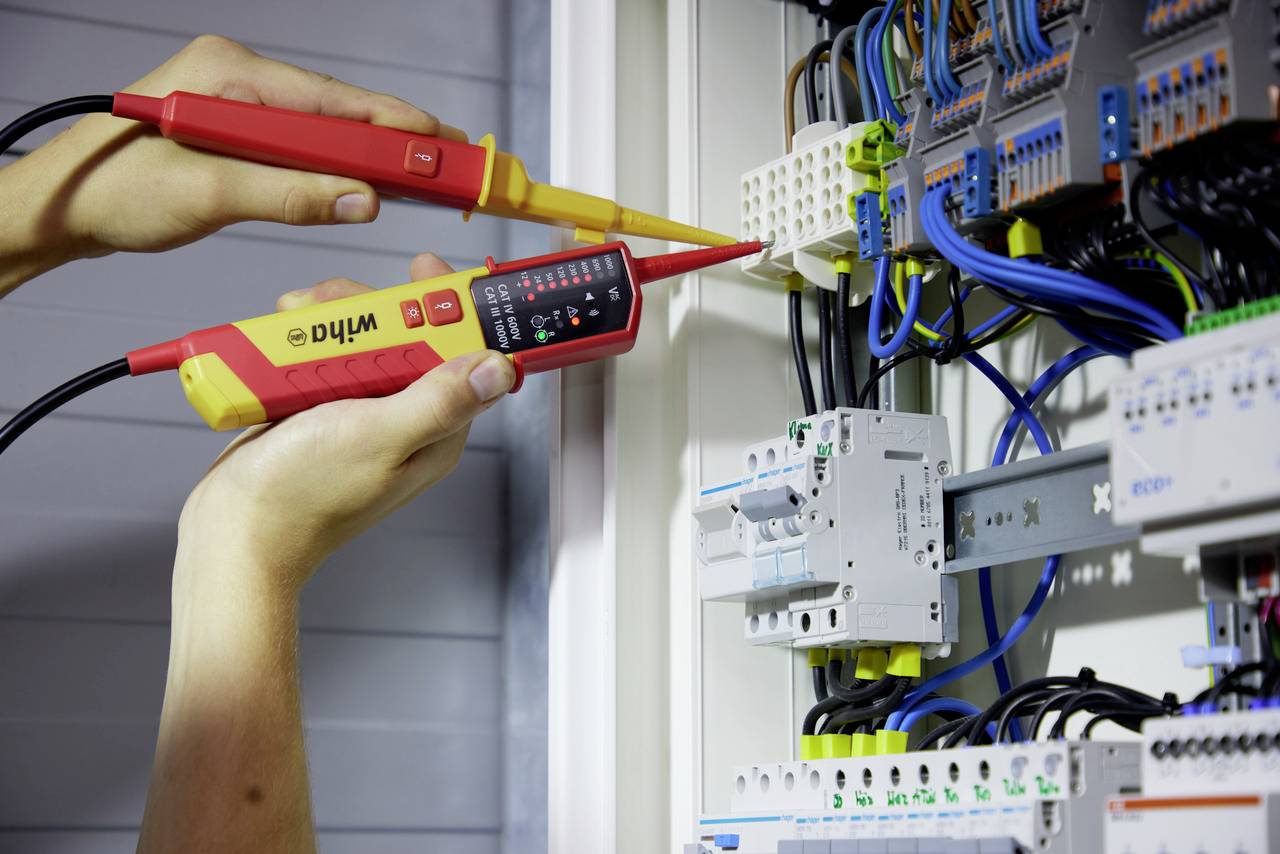 Osoba mierzy napięcie w szafce elektrycznej z licznymi kablami i obwodami za pomocą elektrycznego przyrządu pomiarowego.