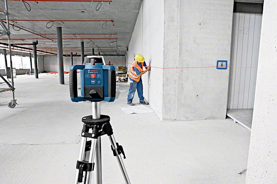 Robotnik budowlany używa laserowego dalmierza do pomiaru odległości w niedokończonym budynku, z urządzeniem na statywie w pierwszym planie.