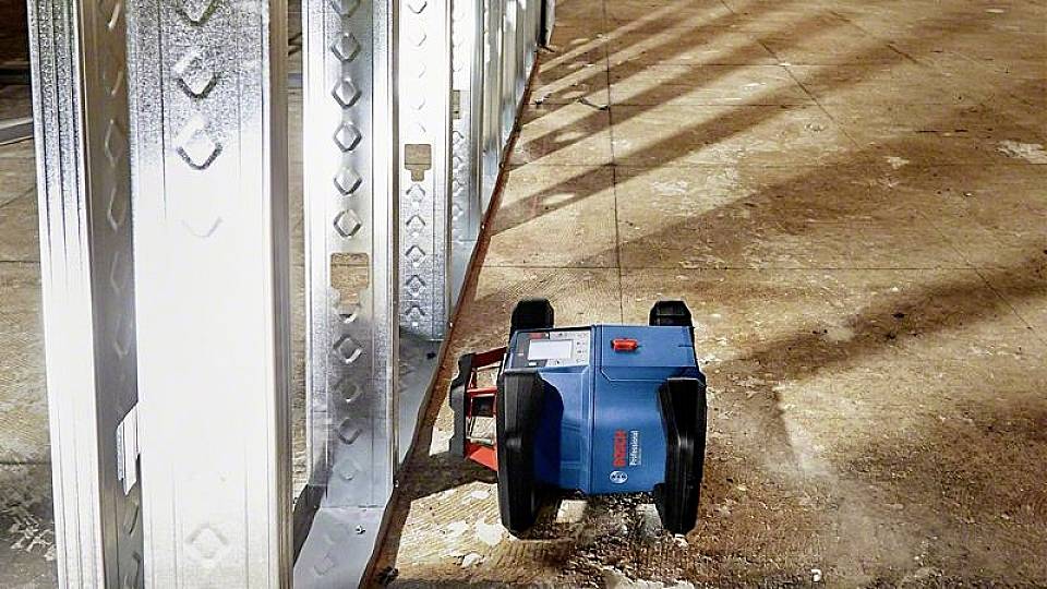 Niebieski robot porusza się wzdłuż metalowej ściany na terenie budowy, prawdopodobnie w celu dokonania pomiaru lub przeprowadzenia inspekcji.