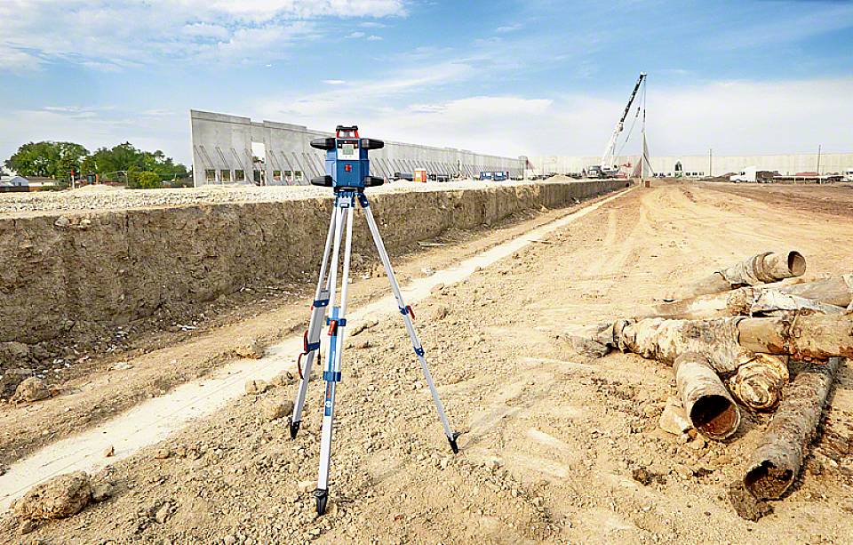 Plac budowy z urządzeniem pomiarowym na pierwszym planie i pracami budowlanymi w tle, w tym żurawiami budowlanymi i rurami.
