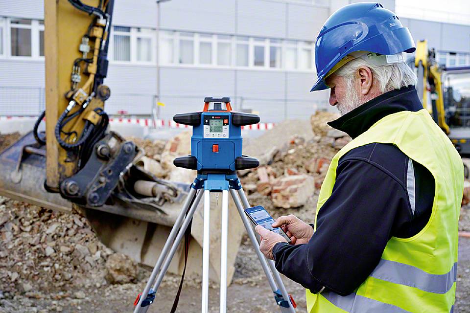 Robotnik budowlany w kasku ochronnym i kamizelce ostrzegawczej obsługuje urządzenie pomiarowe na placu budowy, w tle widoczny jest koparka.