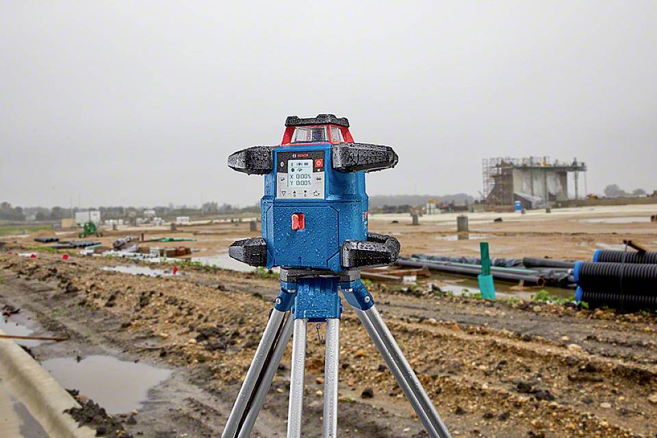 Niebieskie urządzenie geodezyjne na statywie w pierwszym planie na placu budowy; rozmyty tło ukazuje rury i rusztowania.