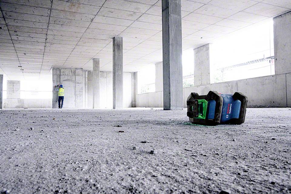 Plac budowy betonowy z wieloma słupami, robotnik budowlany w tle, w przedniej części zielone urządzenie leżące na ziemi.