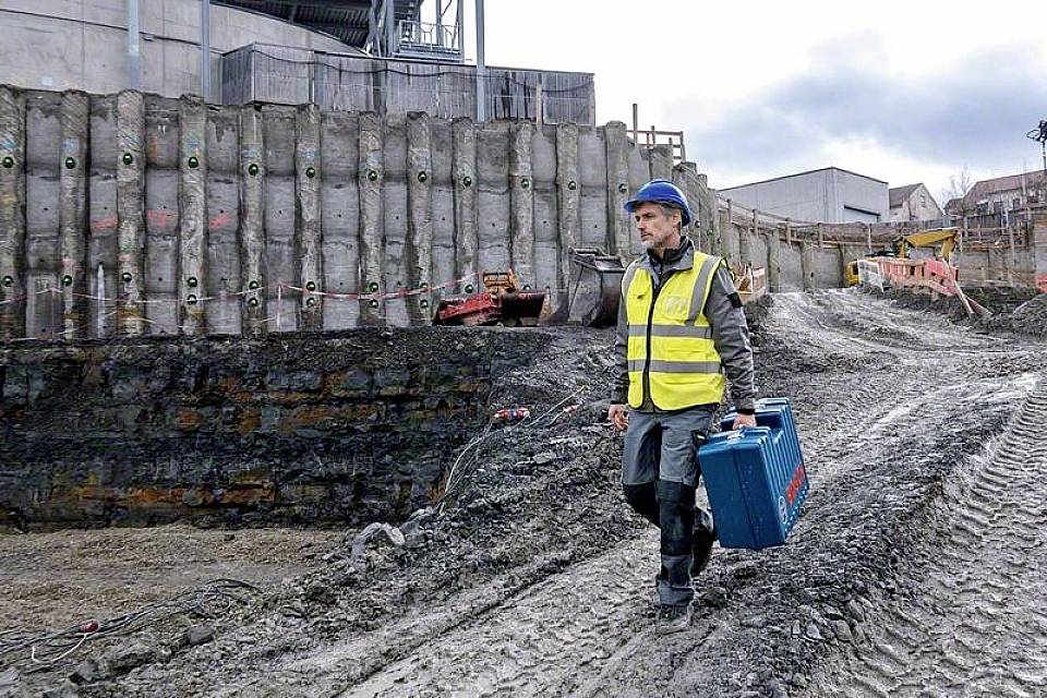 Robotnik budowlany w kasku i kamizelce odblaskowej przechodzi przez plac budowy, trzymając w ręku skrzynkę z narzędziami. W tle widoczne są rusztowania i maszyny budowlane.