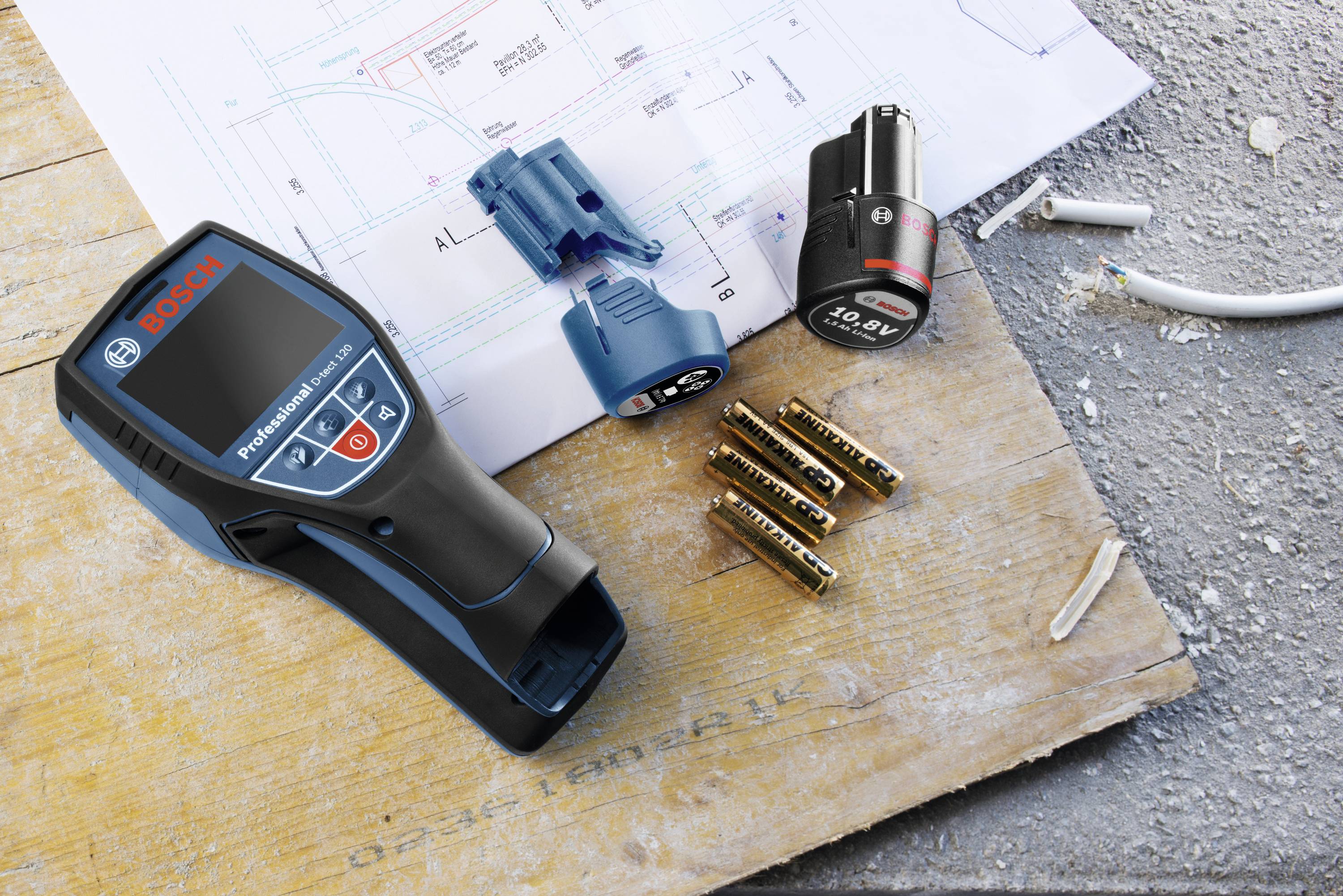 Cyfrowy przyrząd pomiarowy leży na budowie obok planów, baterii i kabli. Wygląda na to, że jest używany do pomiarów technicznych.