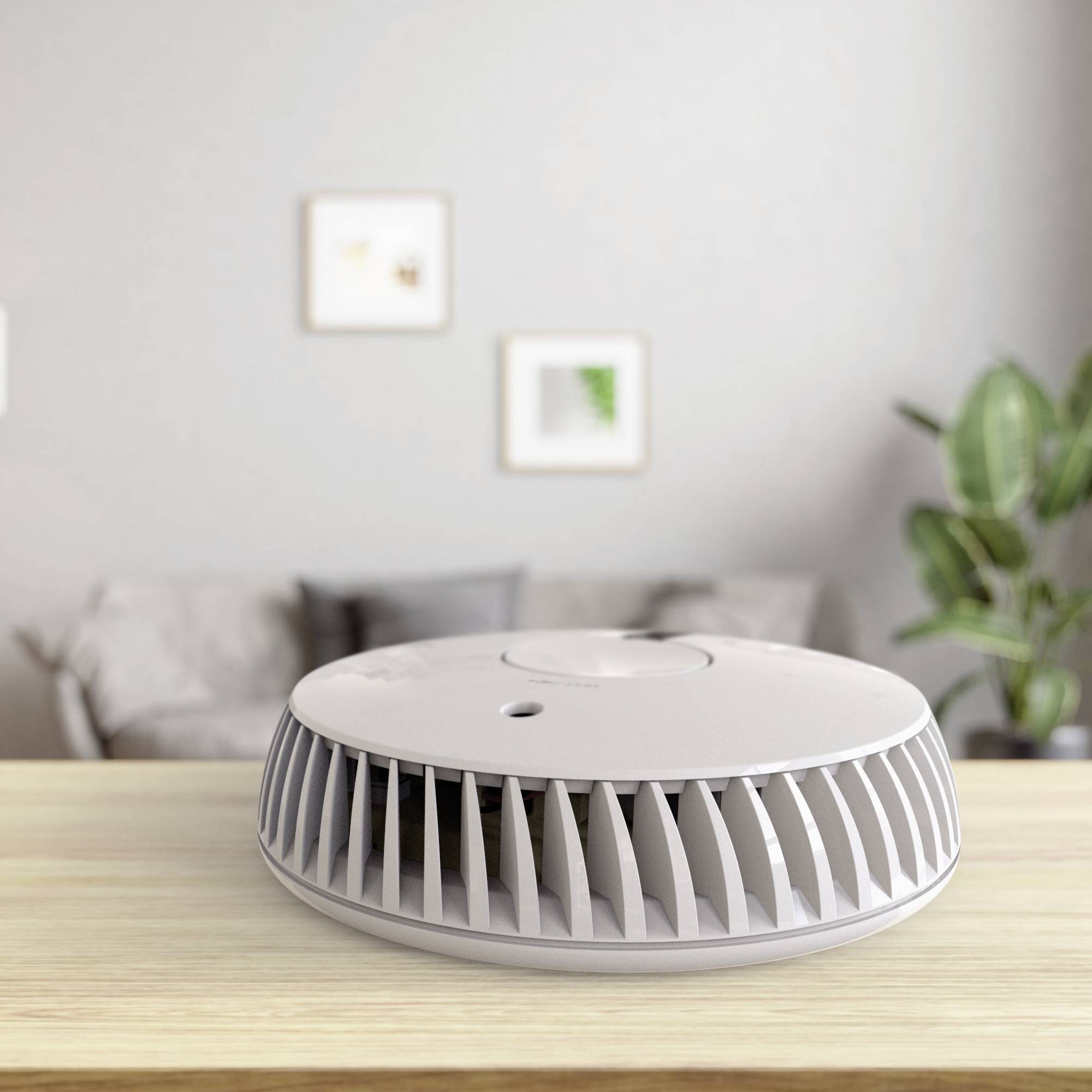 Mały, okrągły oczyszczacz powietrza stoi na drewnianym stole. W tle widać kanapę, rośliny oraz obrazy wiszące na ścianie.