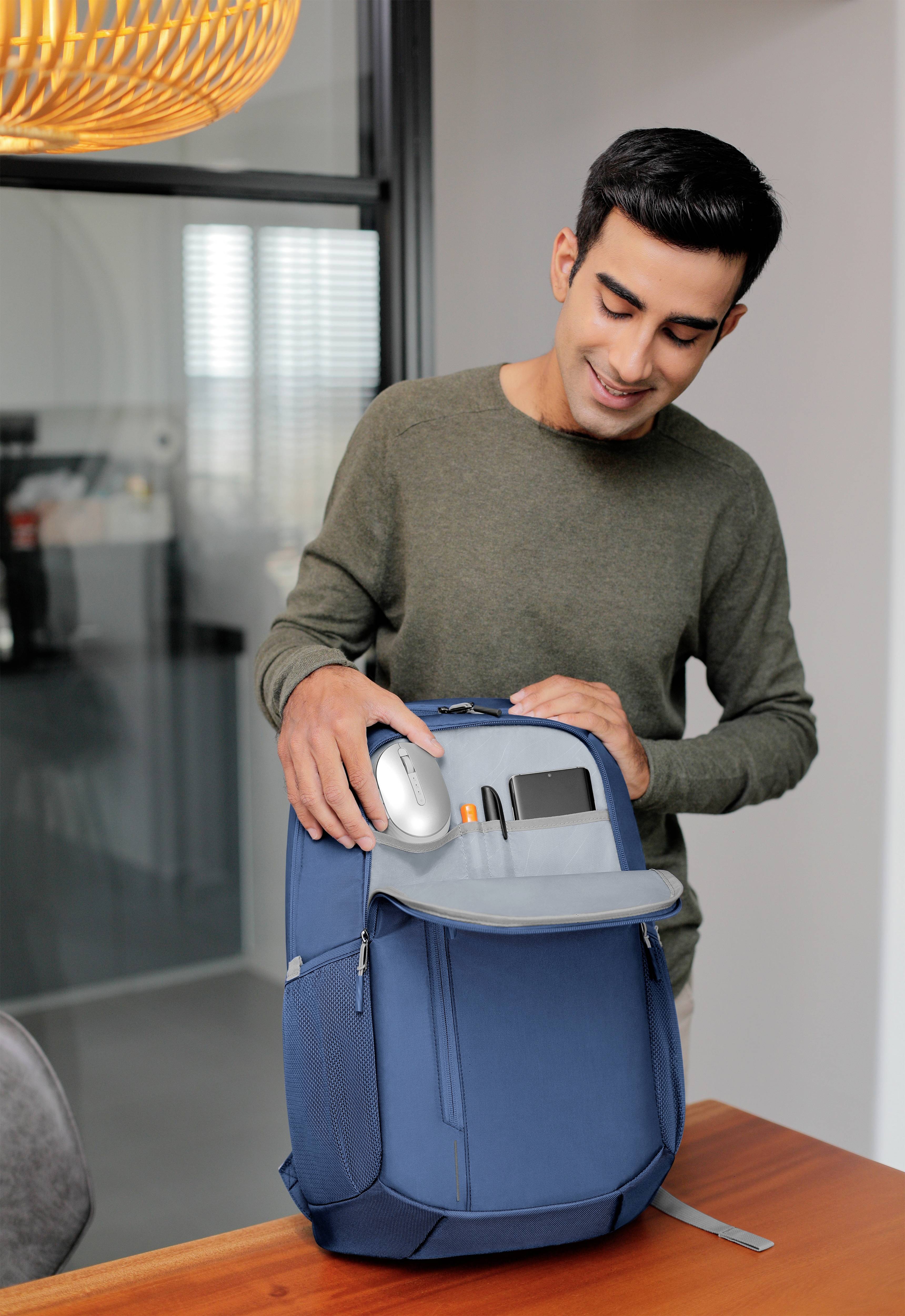 Mężczyzna stoi przy stole i uśmiecha się, otwierając przednią kieszeń niebieskiego plecaka. W tle widać nowocześnie urządzony pokój.