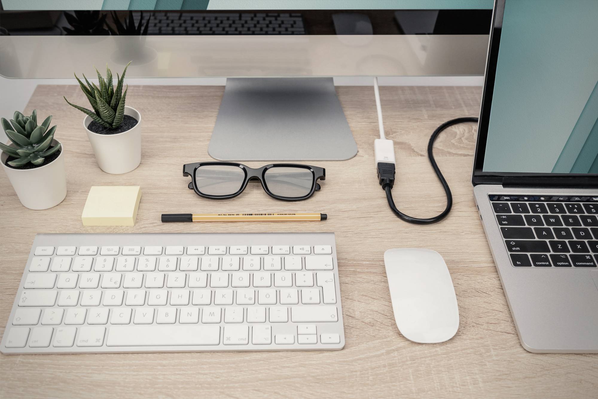 Porządnie zorganizowane biurko z laptopem, klawiaturą, myszą, okularami i dwoma małymi roślinami w doniczkach. Kabel łączy laptopa z monitorem.