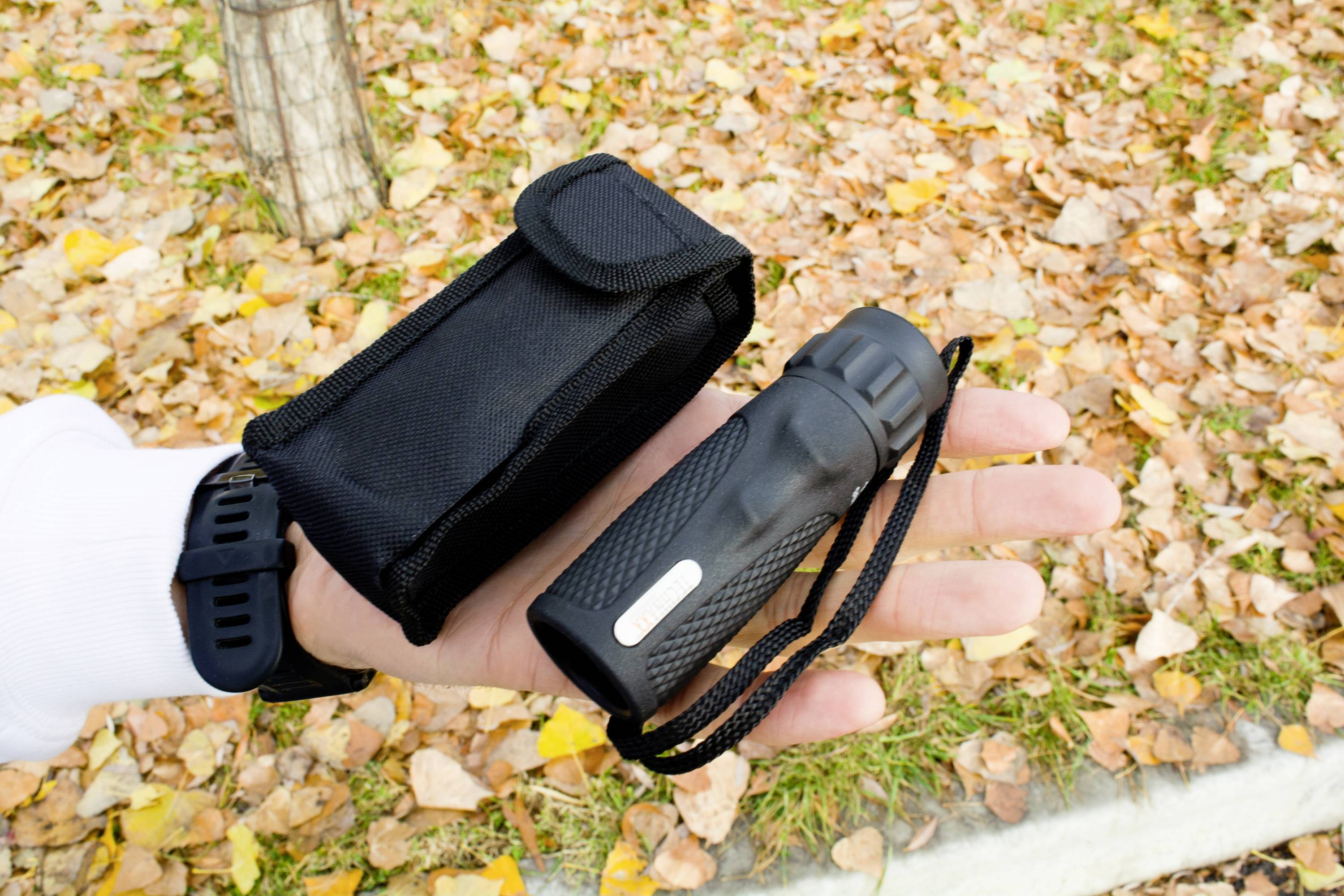 Osoba trzyma czarną latarkę i pasujące do niej etui. W tle widoczne są jesienne liście leżące na ziemi.