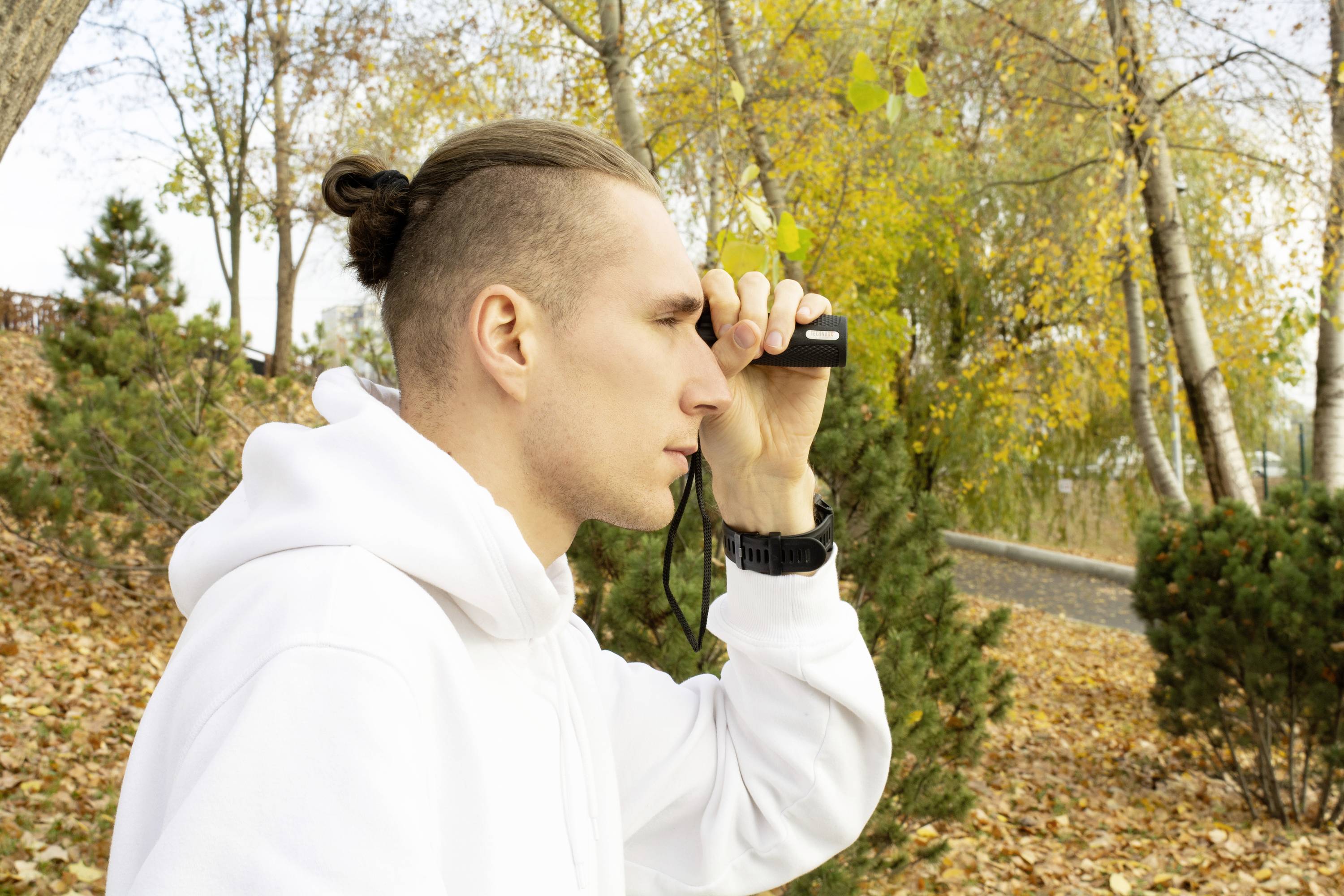 Mężczyzna w białej bluzie z kapturem patrzy przez lornetkę w jesiennym parku. W tle drzewa liściaste.