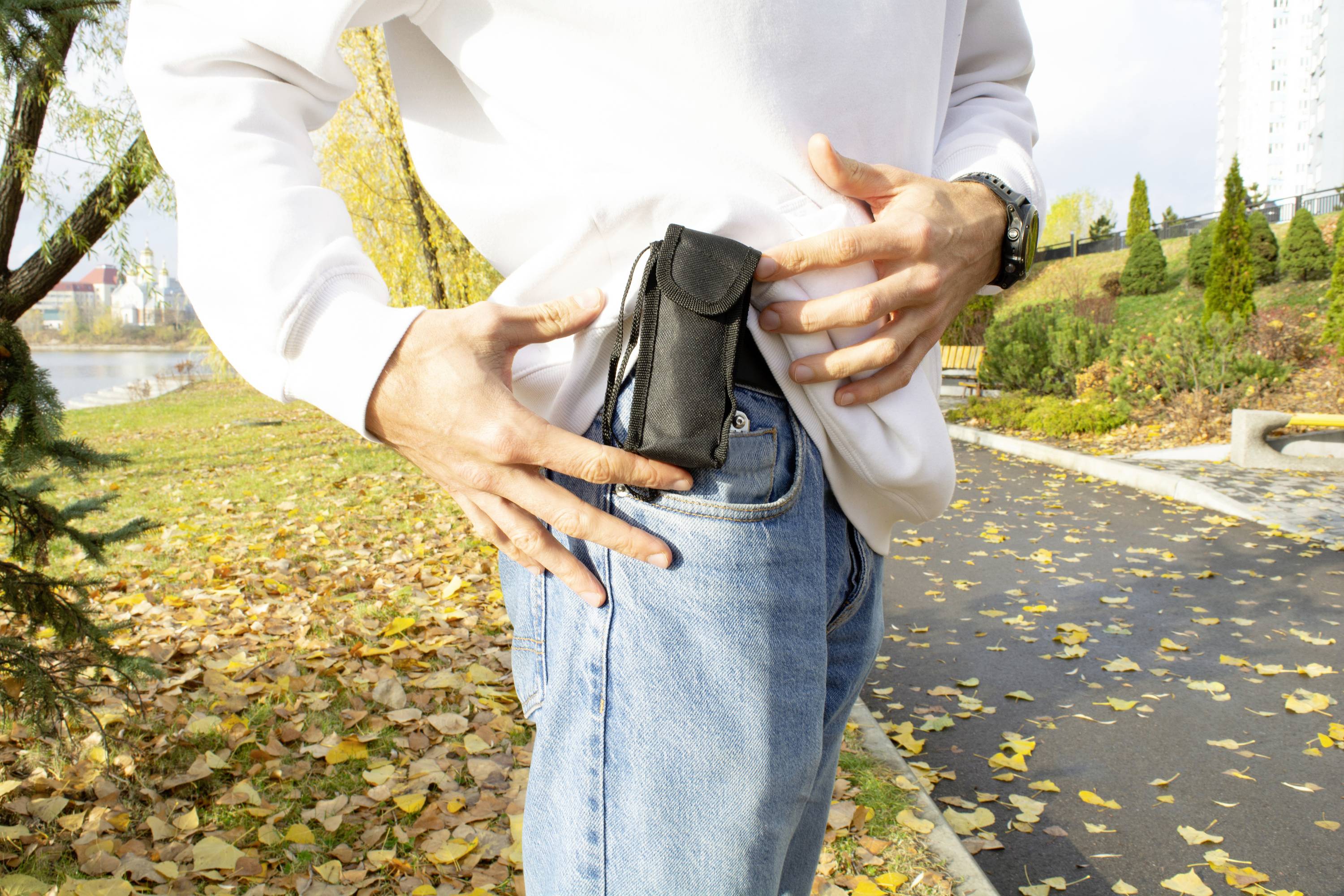Osoba wkłada czarne etui na smartfona do kieszeni spodni. W tle widać jesienny park i chodnik.