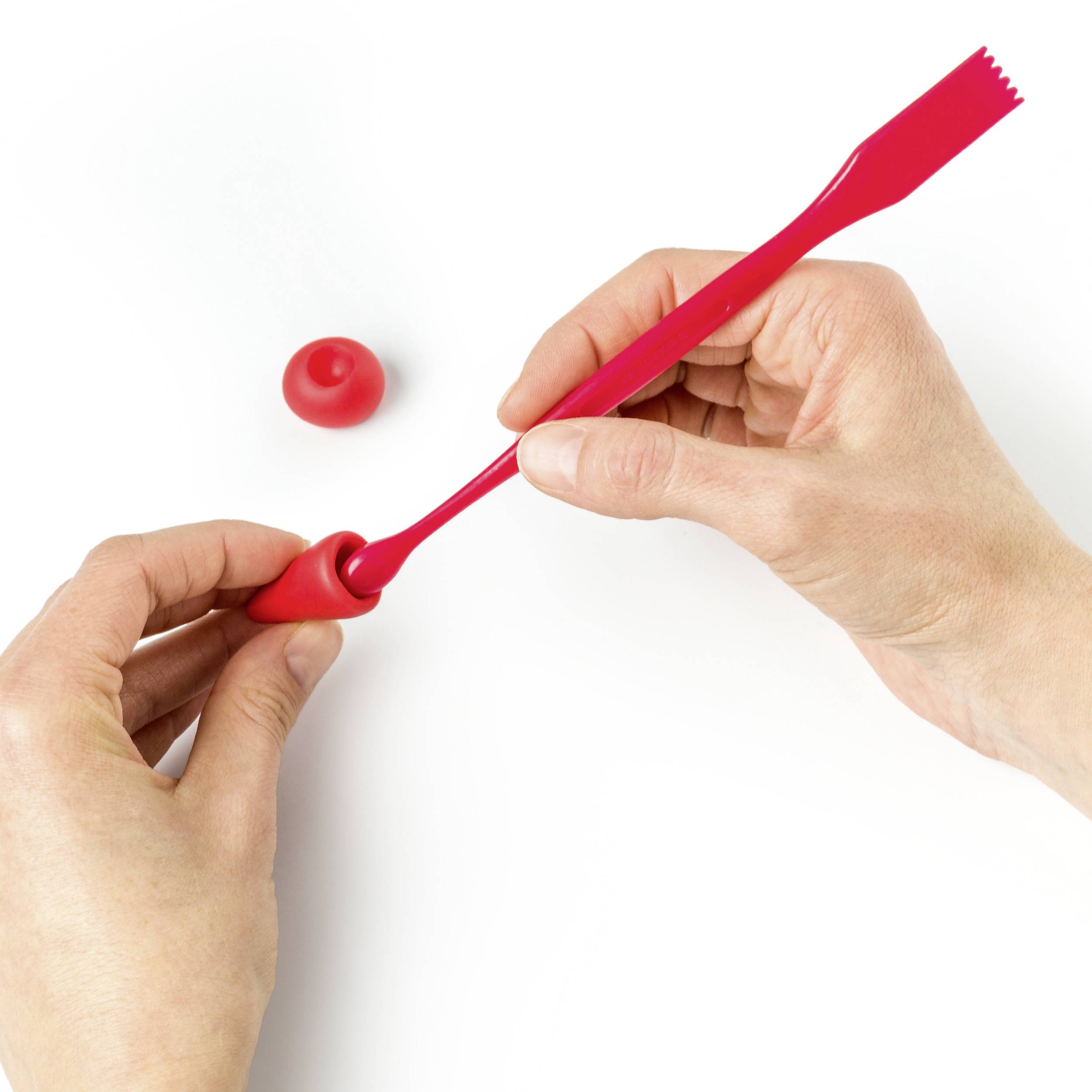 Dwie ręce używają czerwonego narzędzia modelarskiego do obróbki czerwonej kuli glinianej na białej powierzchni.