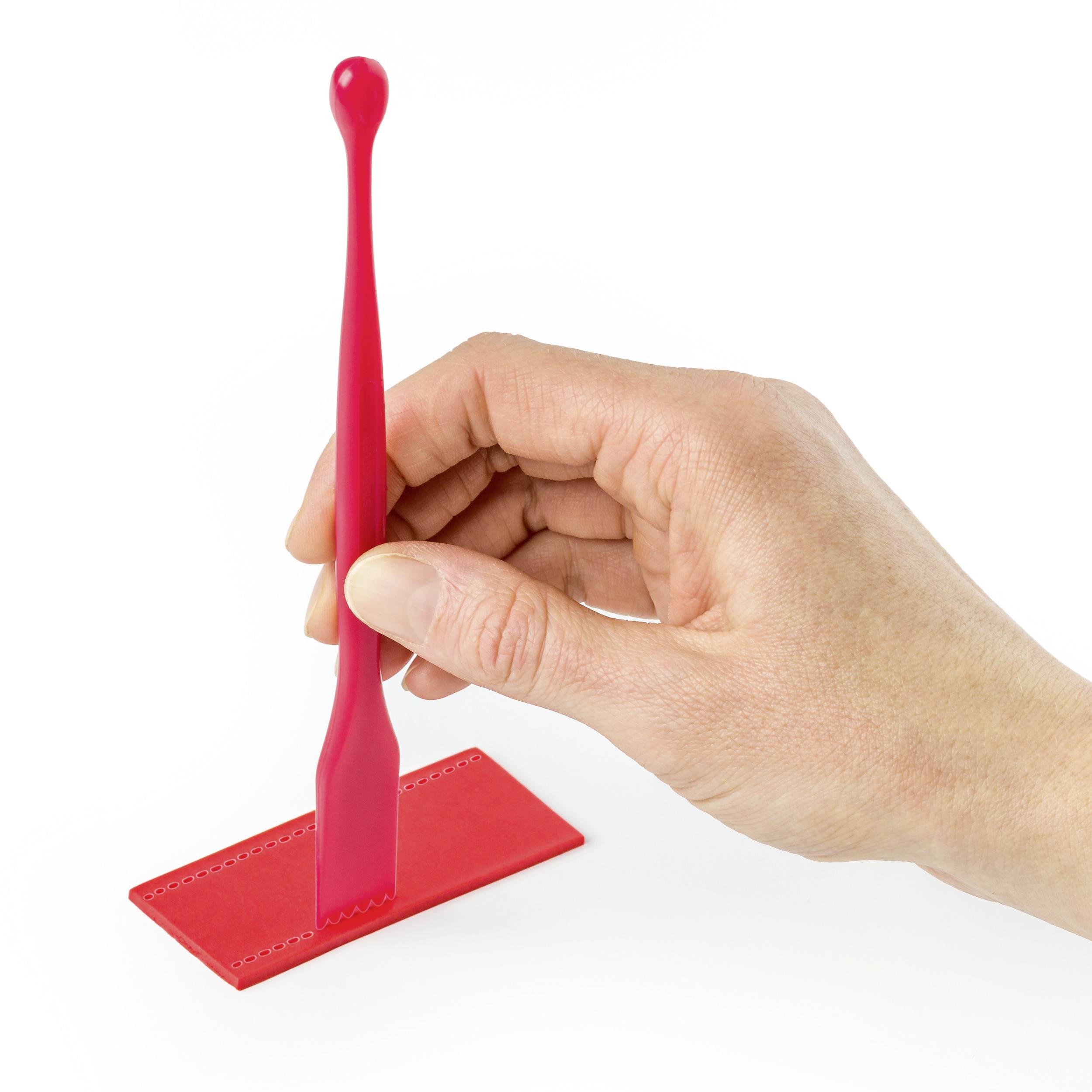 Dłoń trzyma czerwony obiekt designerski, przypominający ręczny zegar słoneczny, umieszczony na płaskiej, czerwonej podstawie.