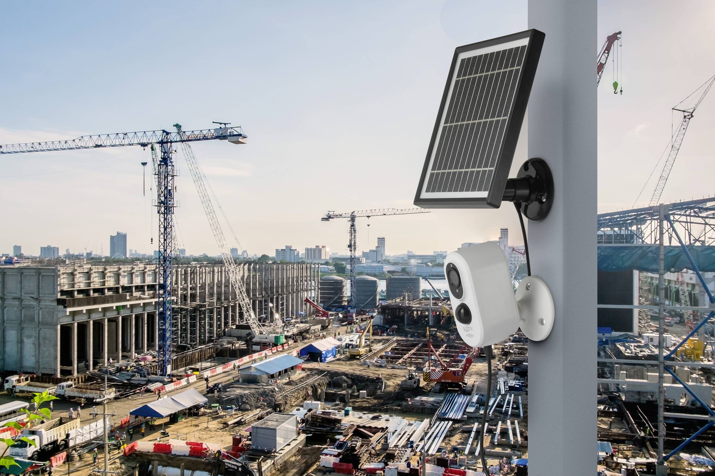 Instalacja monitoringu i solarna na słupie przed rozległym terenem budowy, z dźwigami i strukturą budynków w tle.