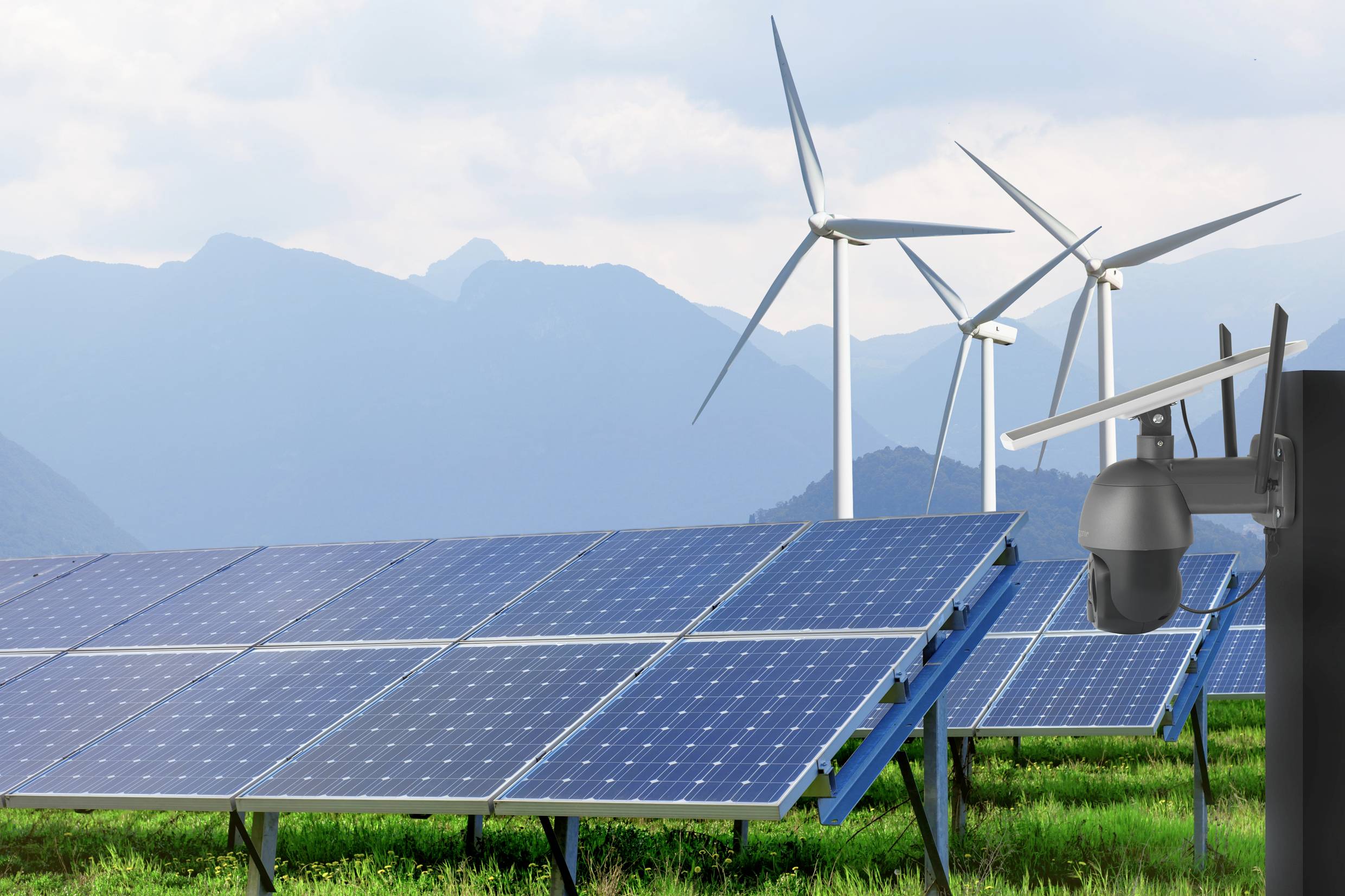 Panele słoneczne na zielonej łące przed górami, z elektrowniami wiatrowymi w tle. Monitoring kamerowy na pierwszym planie. Temat: energia odnawialna.