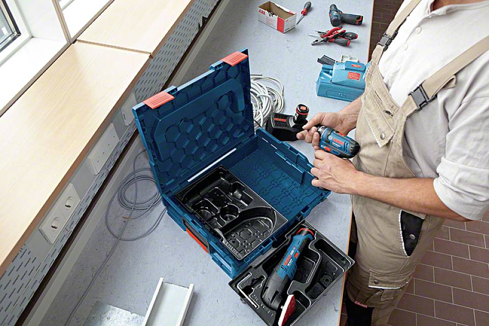 Osoba w stroju roboczym trzyma elektronarzędzie i przygotowuje je do użycia. Na stole leżą różne narzędzia i kable.