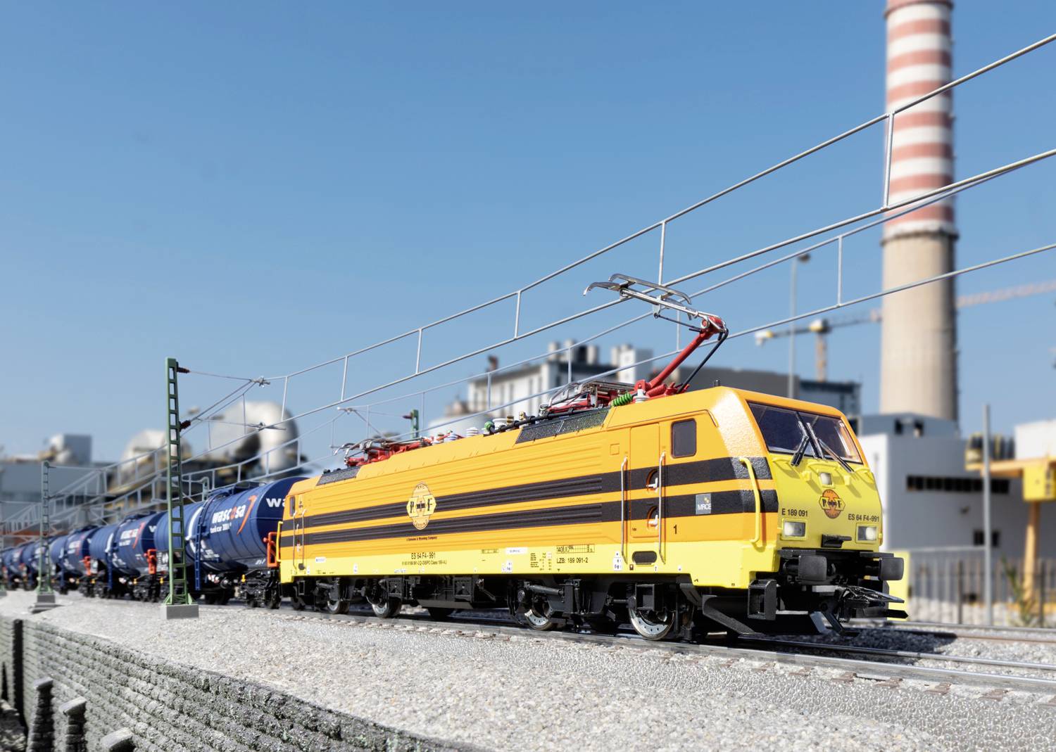 Żółta lokomotywa ciągnie pociąg towarowy z niebieskimi cysternami wzdłuż kompleksu przemysłowego, w tle komin na tle błękitnego nieba.