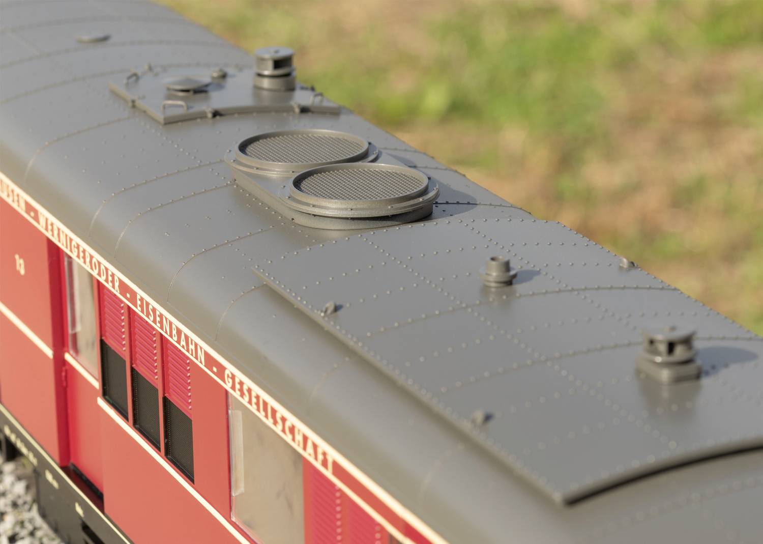 Zbliżenie czerwonego wagonu kolejki modelowej z szarym dachem i otworami wentylacyjnymi. W tle rozmyte trawy.