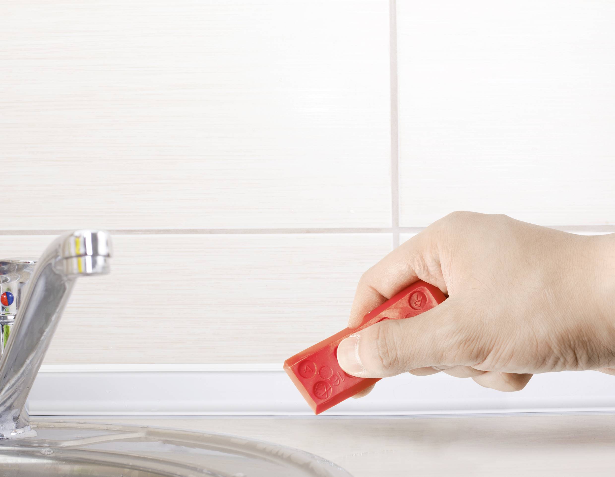 Ręka trzyma czerwoną klocką Lego nad zlewem na tle białych kafelków.