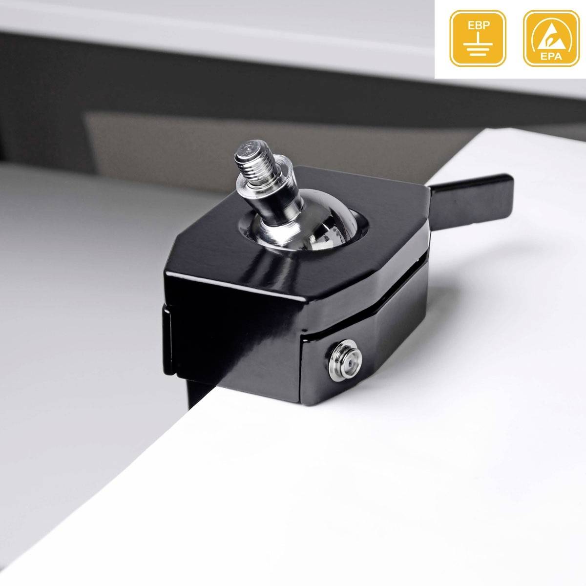 Czarny metalowy przyrząd jest zamocowany na krawędzi stołu. Posiada regulowaną dźwignię i służy do uziemiania elektroniki.