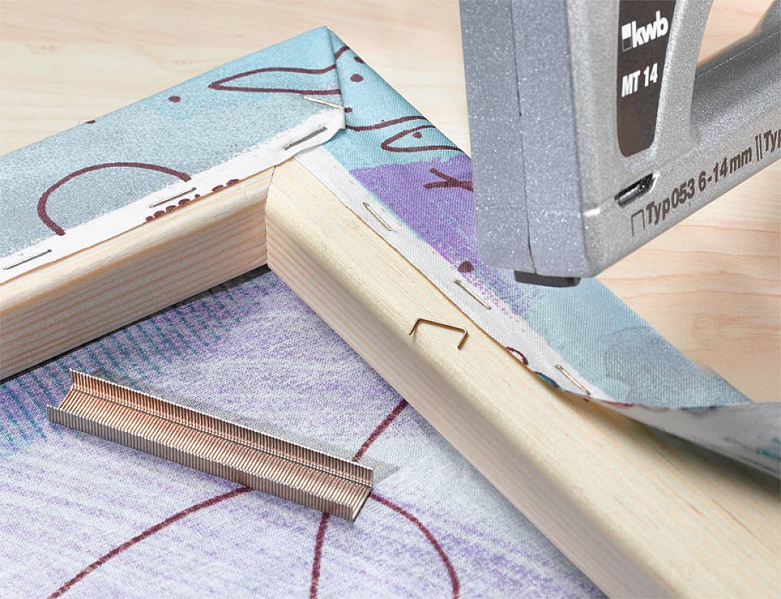 Pistolet tapicerski mocuje płótno do drewnianej ramy; obok leży pasek zszywek.