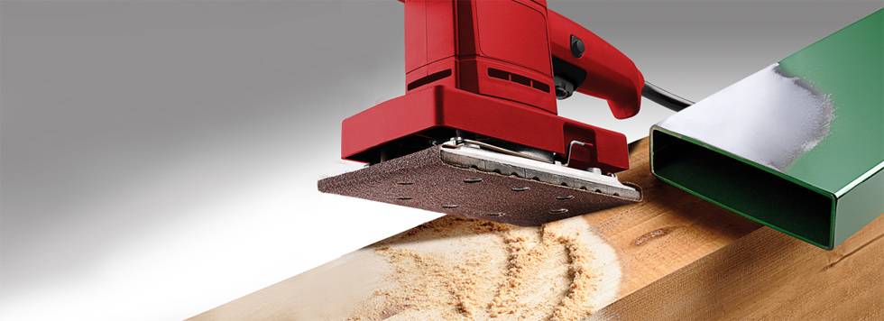 Czerwona szlifierka elektryczna szlifuje powierzchnię drewna. Po jej prawej stronie leży zielony kawałek metalu.