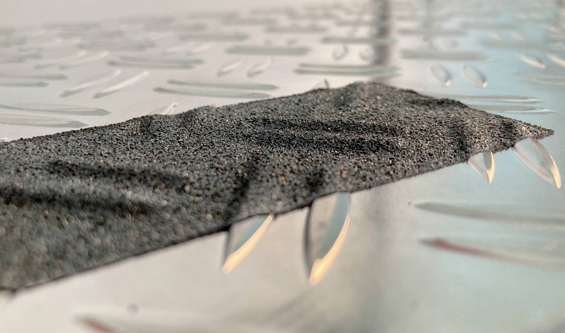 Zbliżenie na prostokątny, antypoślizgowy pasek na metalowej powierzchni. Pasek ma chropowatą teksturę.