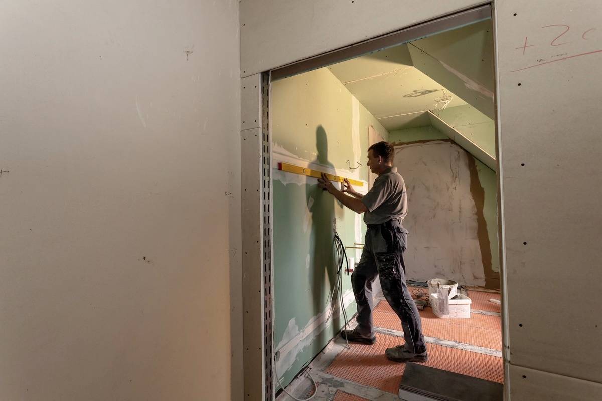 Osoba pracuje w pomieszczeniu w trakcie remontu, używając poziomicy przy zielonej ścianie, aby wyrównać lub sprawdzić jej płaszczyznę.