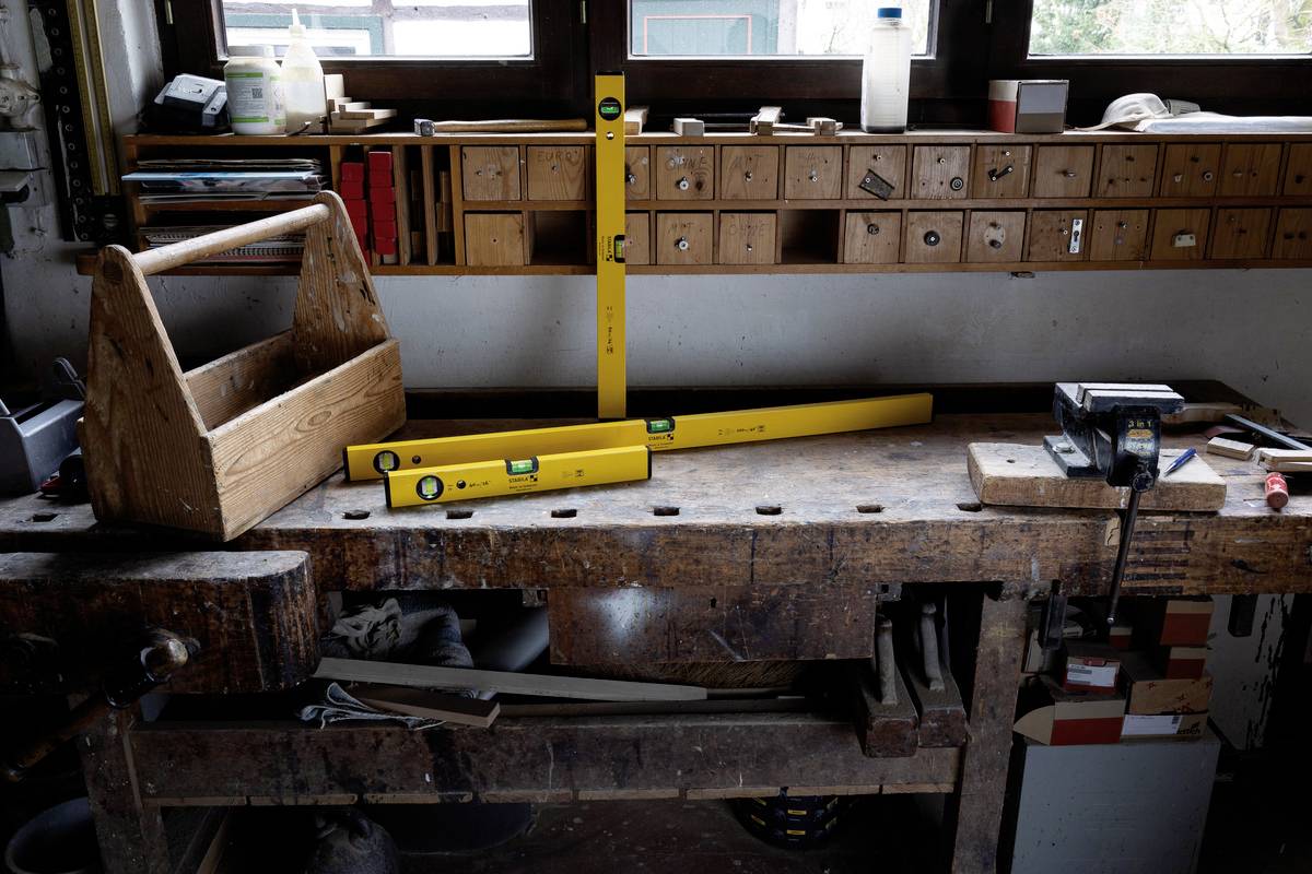 Stół warsztatowy w warsztacie z żółtymi poziomicami, skrzynką narzędziową i imadłem. Drewniane szuflady w tle.