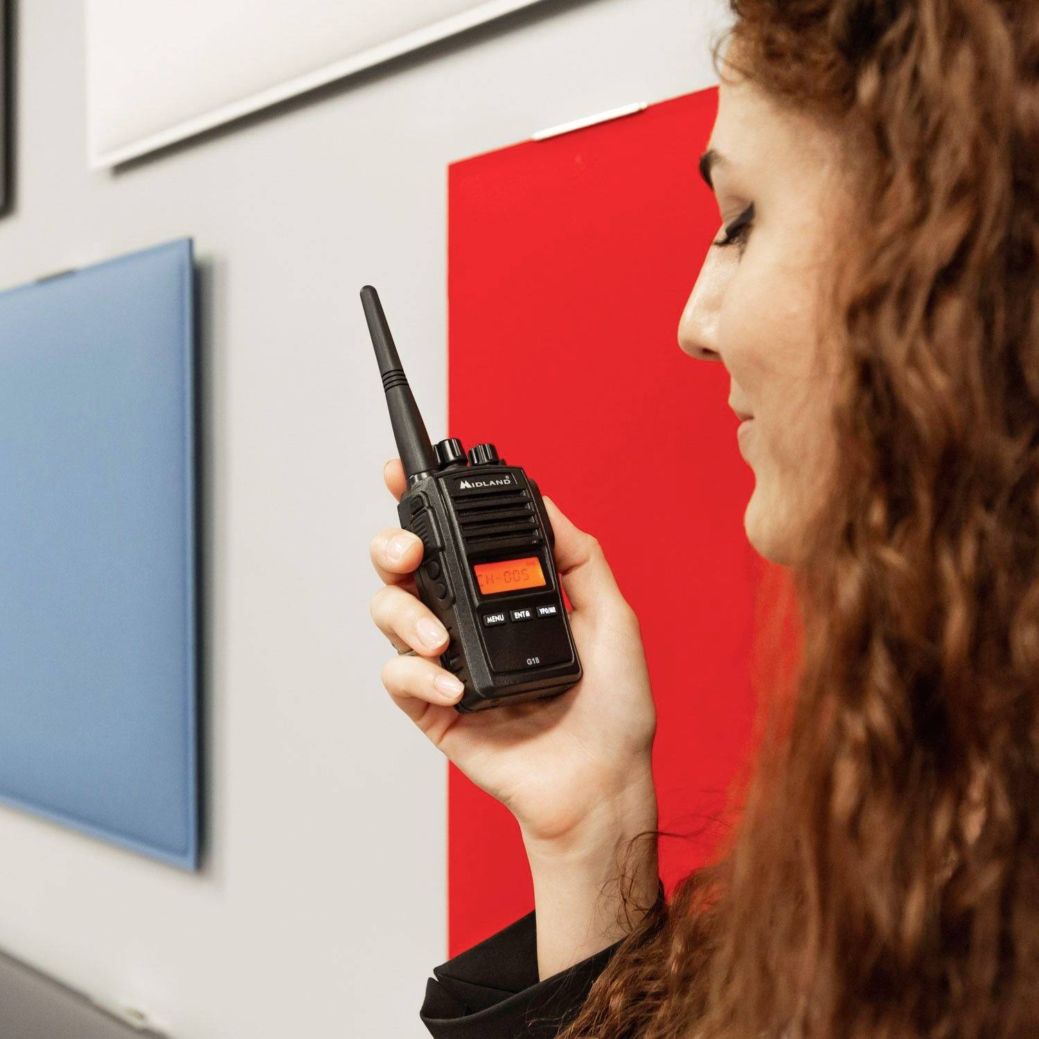 Osoba trzyma radiotelefon, gotowa do jego użycia. Na tle widoczne są kolorowe tablice na ścianie.