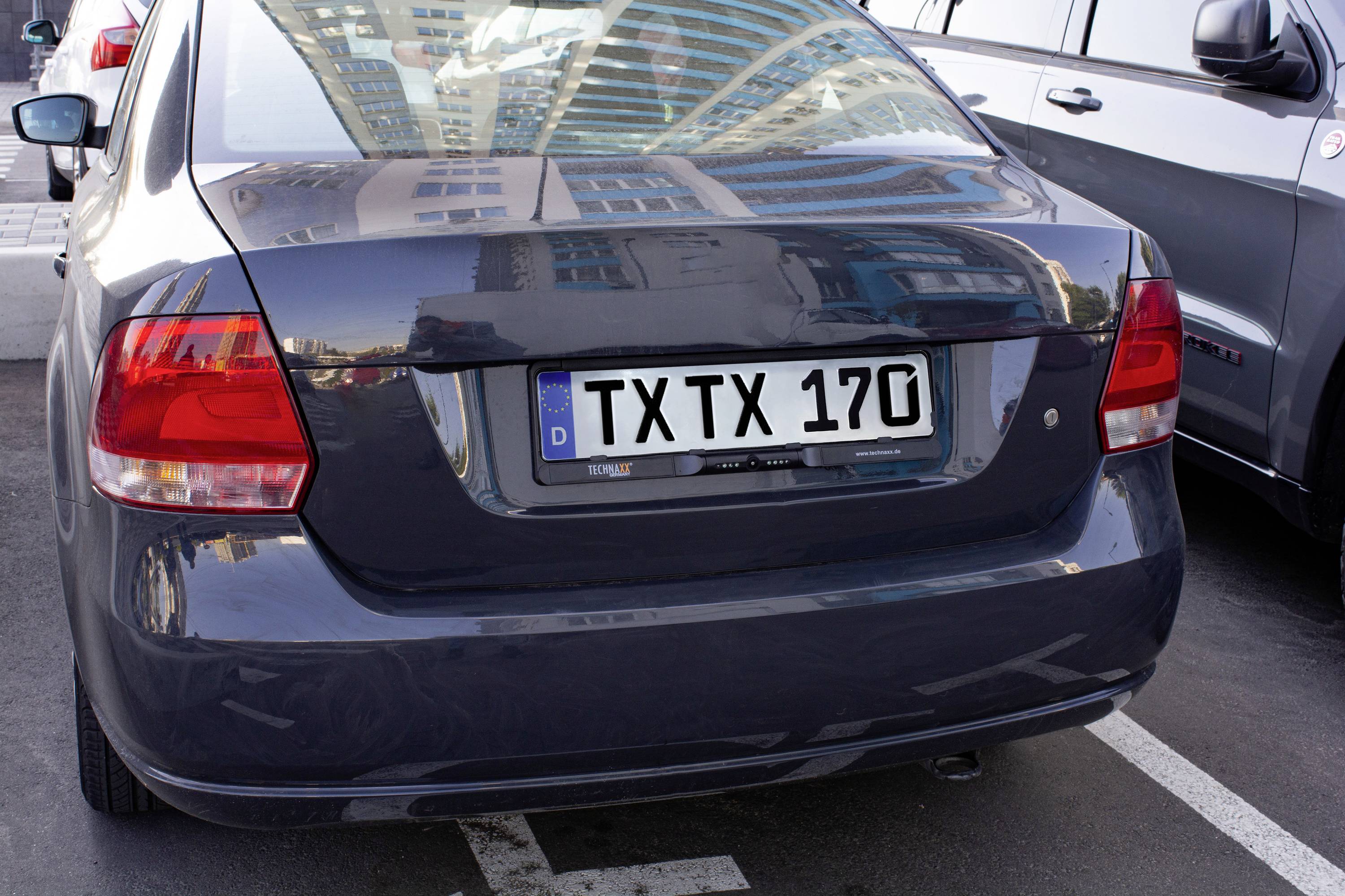 Ciemny samochód stoi na parkingu; numer rejestracyjny to 'TXTX 170'. W tle widać wysokie budynki.