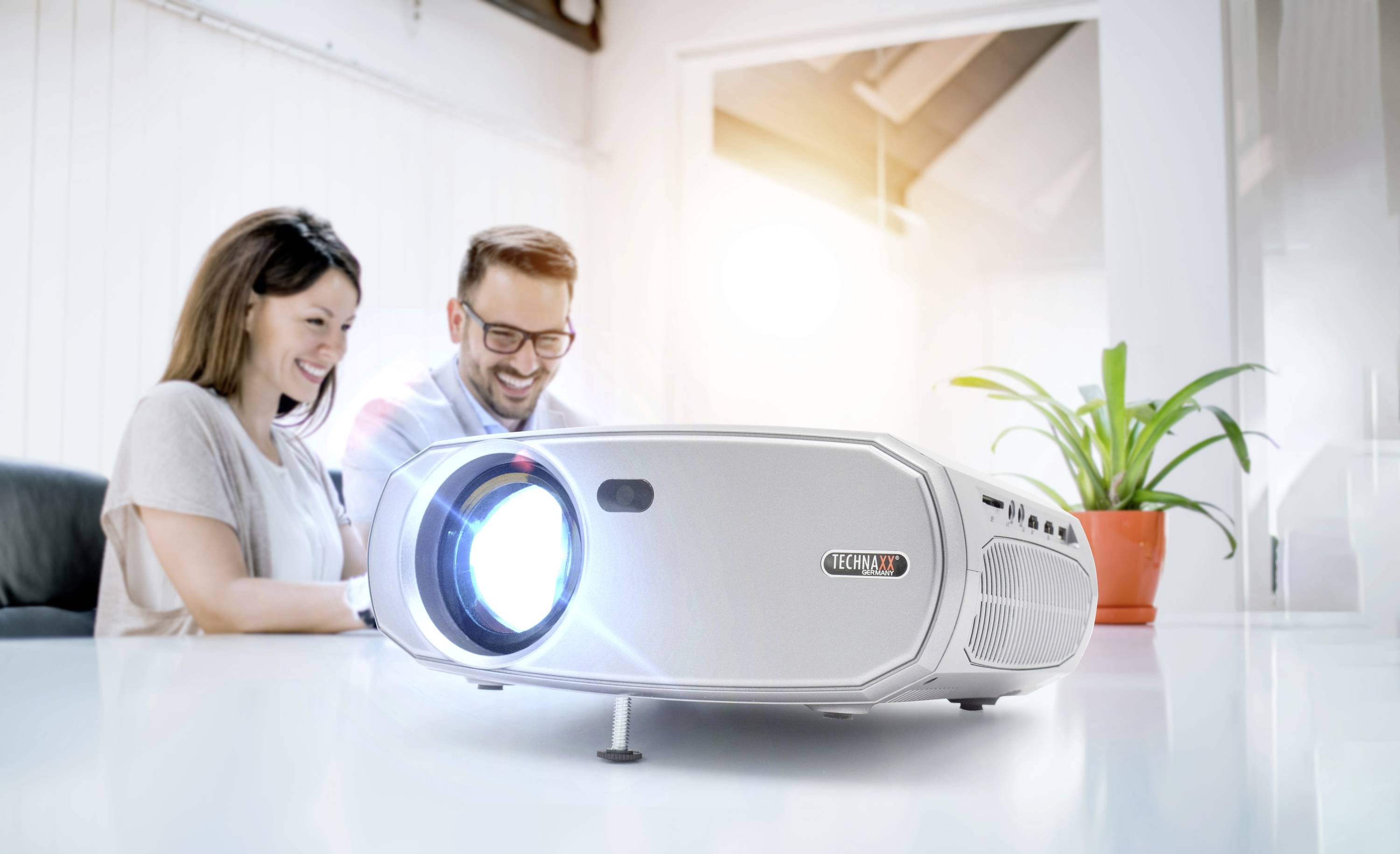 Projektor rzuca jasne światło na stół. W tle dwie osoby siedzą uśmiechnięte obok siebie podczas spotkania.