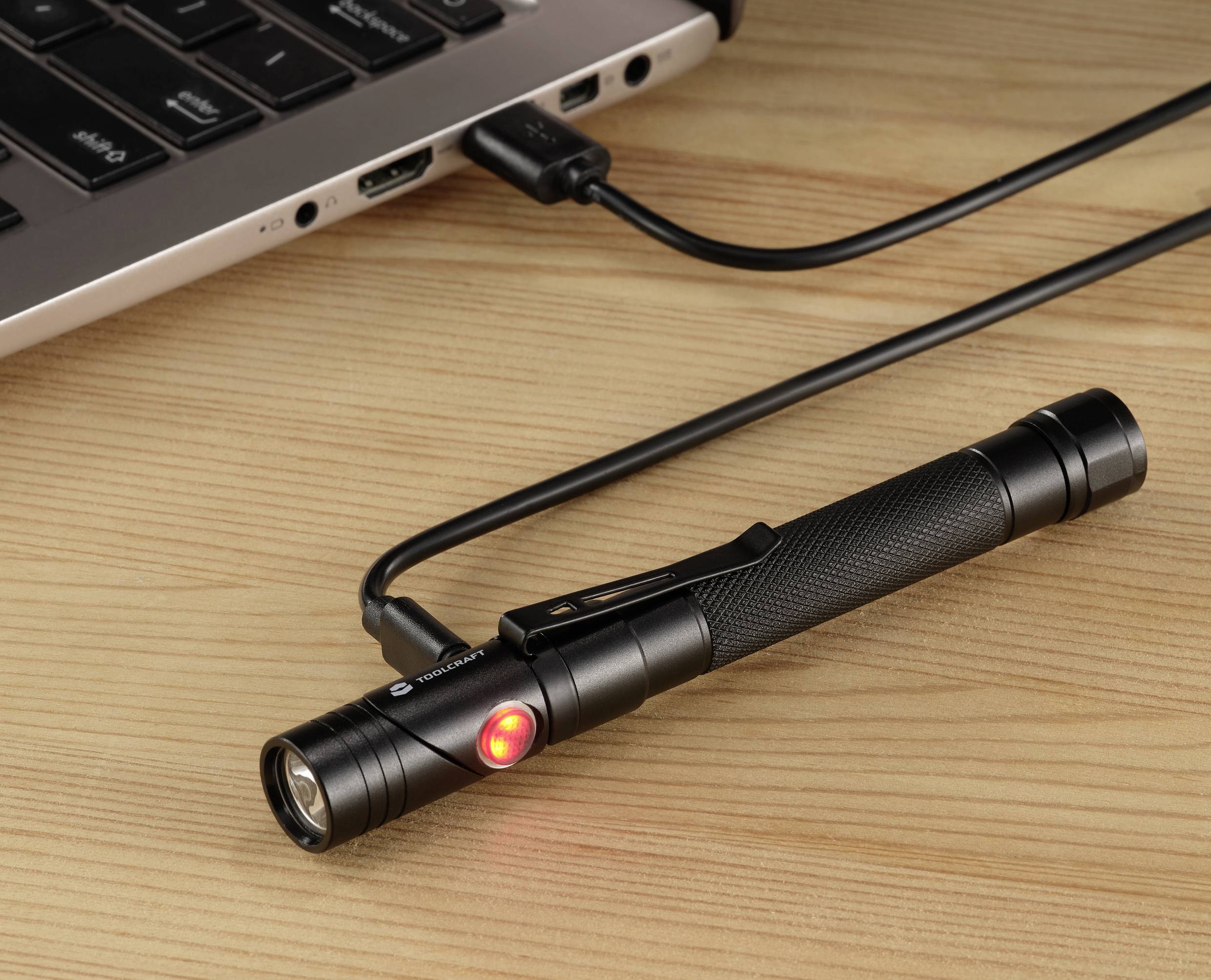 Czarna latarka leży na drewniano-brązowym stole, podłączona do laptopa za pomocą kabla USB, czerwone światło na latarce świeci.