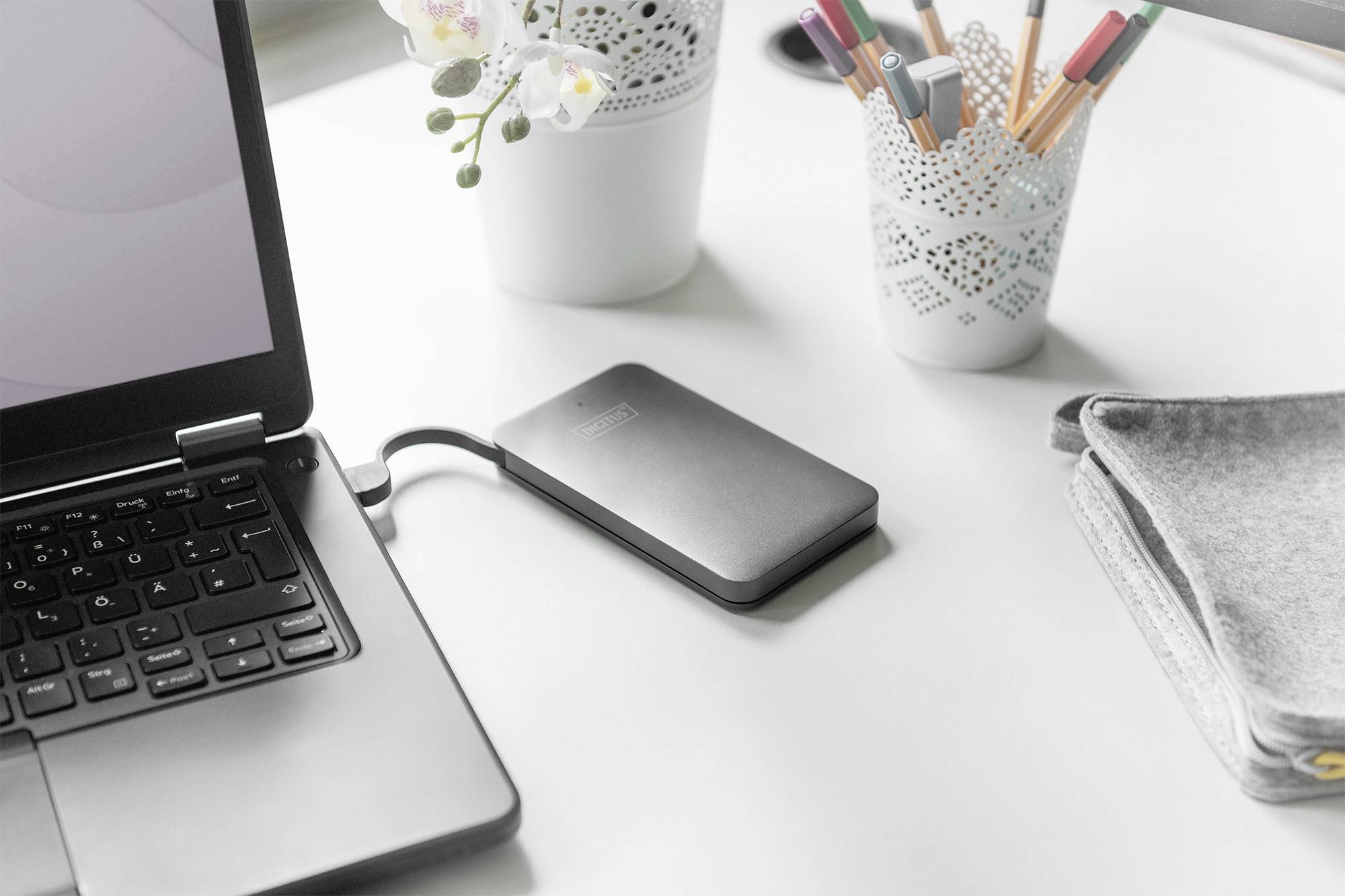 Zewnętrzny dysk twardy podłączony do laptopa na biurku, w tle długopisy i rośliny.