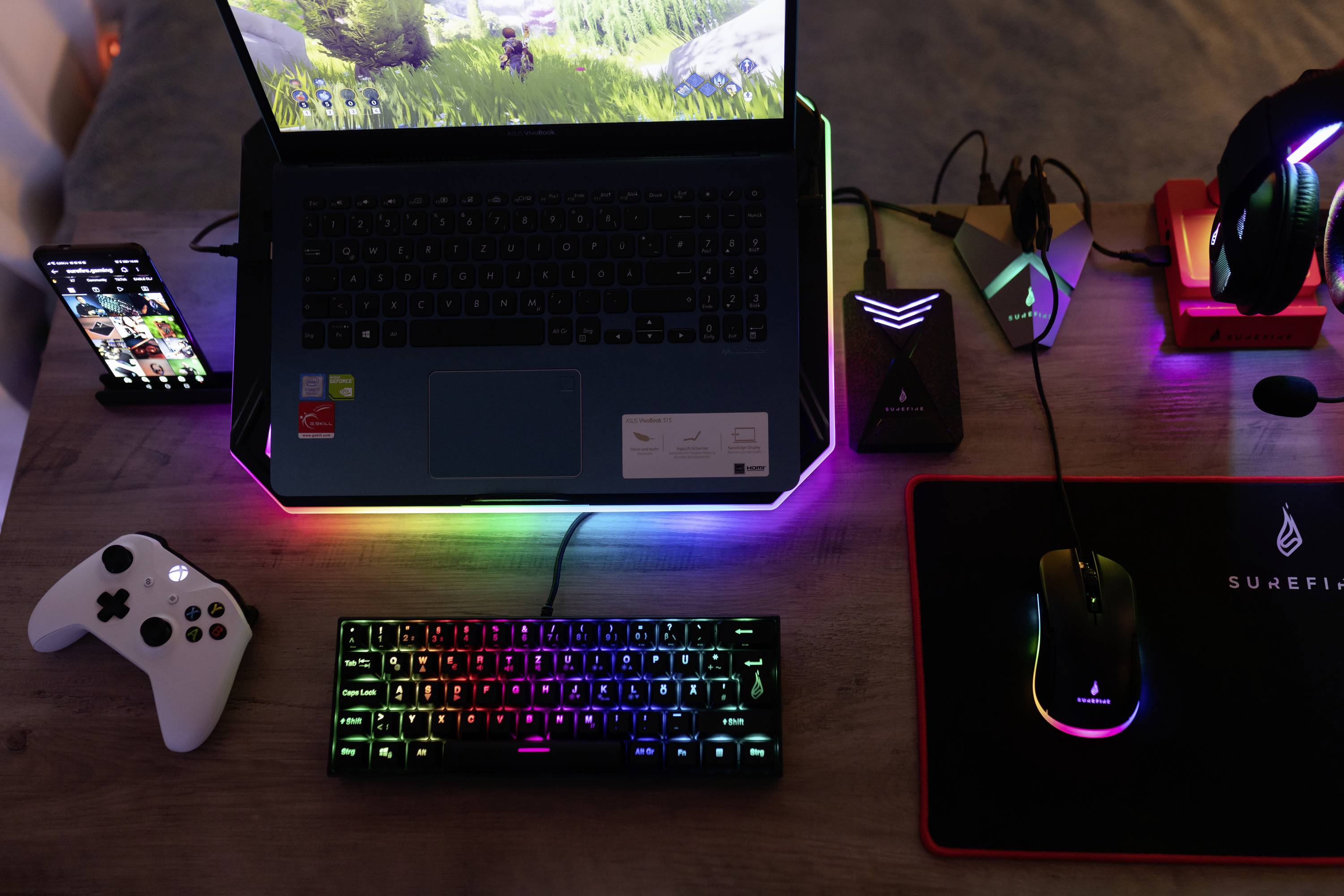 Biurko z podświetleniem LED zmieniającym kolory prezentuje laptopa, klawiaturę, mysz, smartfona i kontroler, wszystkie z podświetlanymi akcentami.