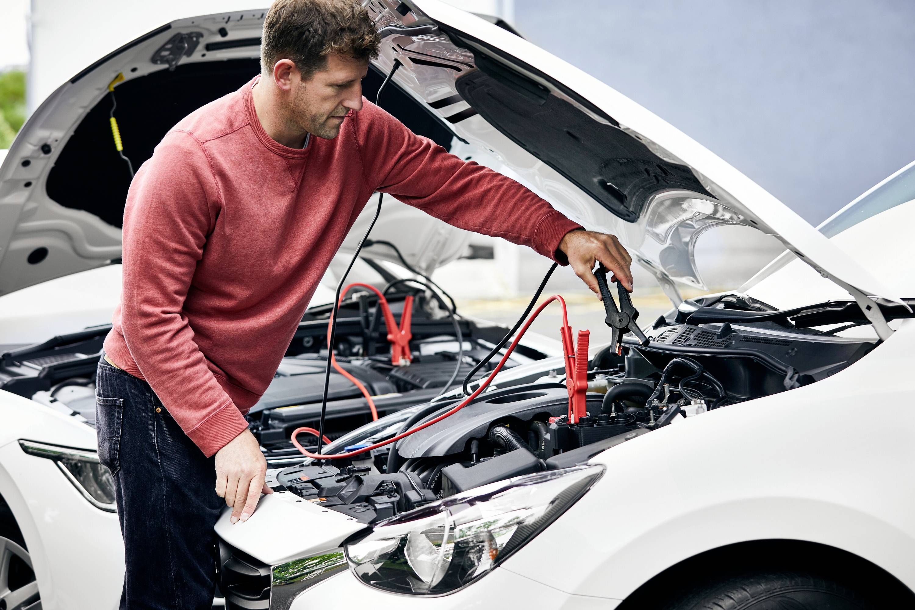 Mężczyzna w czerwonym swetrze podłącza kable rozruchowe do samochodu, z otwartą maską silnika, na parkingu.