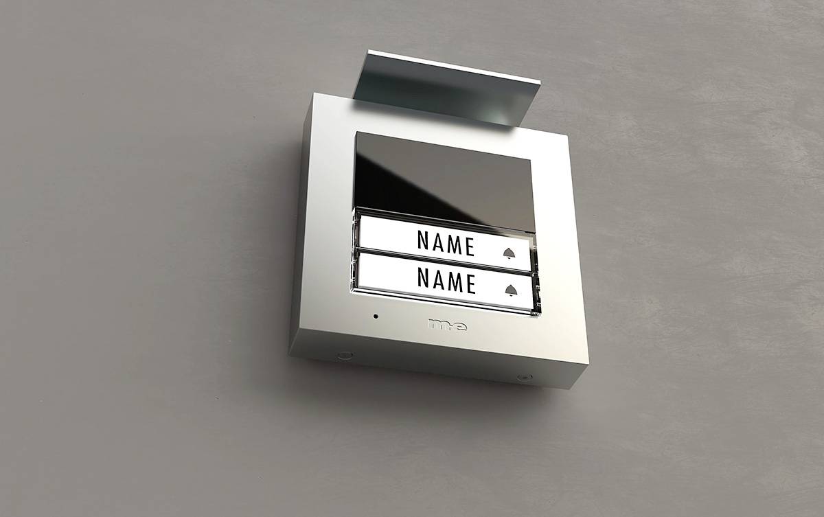 Podwójny przycisk dzwonkowy z dwoma tabliczkami z nazwiskami obok siebie, zamontowany na ścianie, z małym daszkiem ochronnym nad nim.