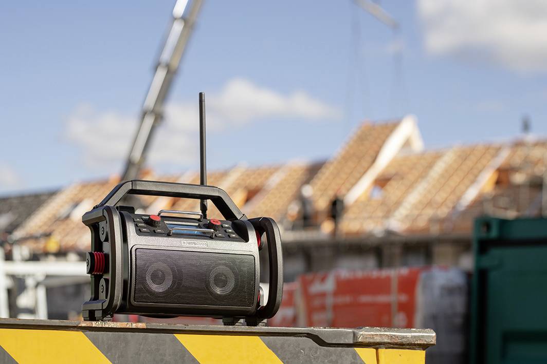 Radio budowlane stoi na skrzyni oznaczonej czarno-żółtymi pasami. W tle widać nieostre prace budowlane.