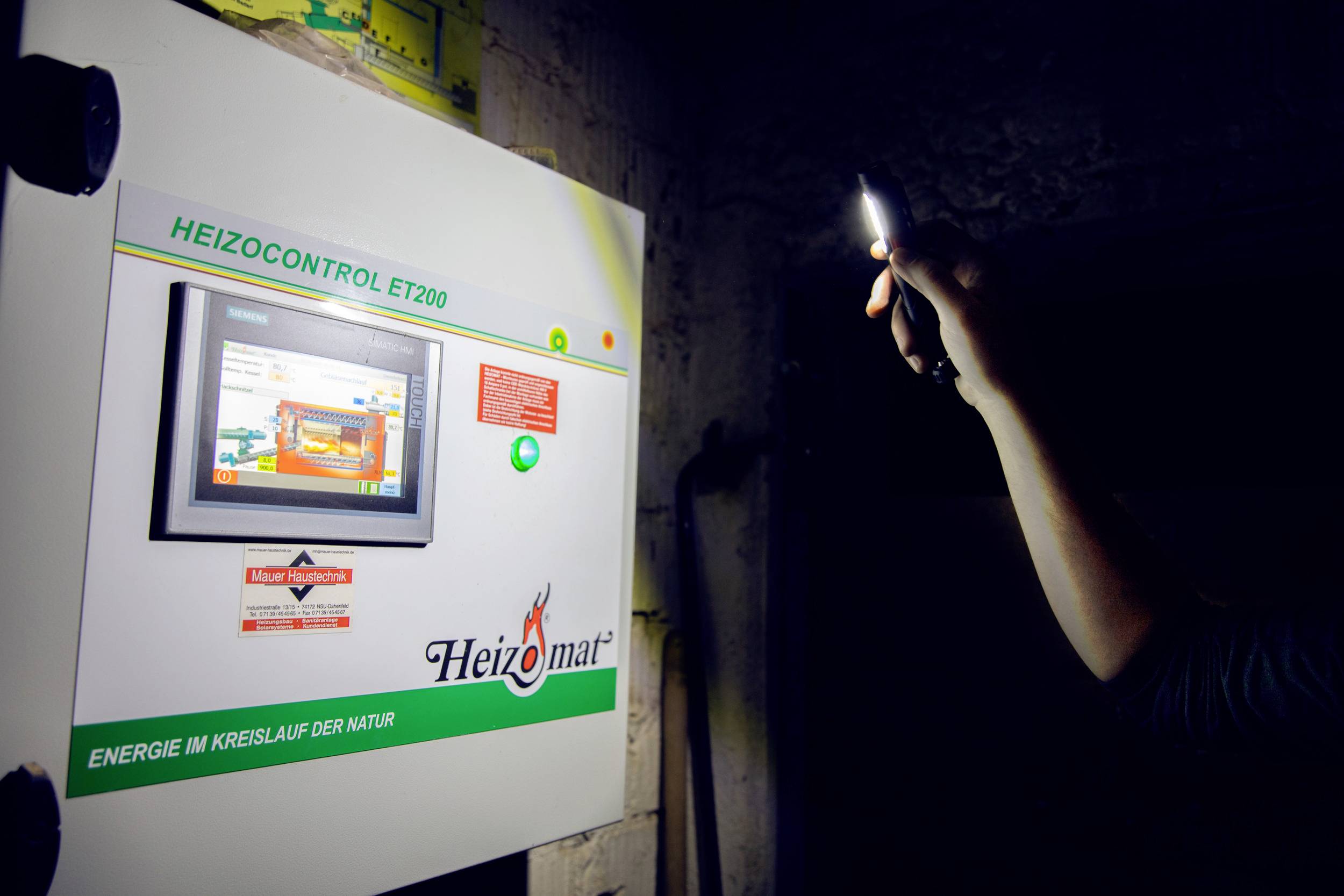 Człowiek oświetla latarką skrzynkę sterowania ogrzewaniem z wyświetlaczem, na którym widnieje napis 'Heizomat'.