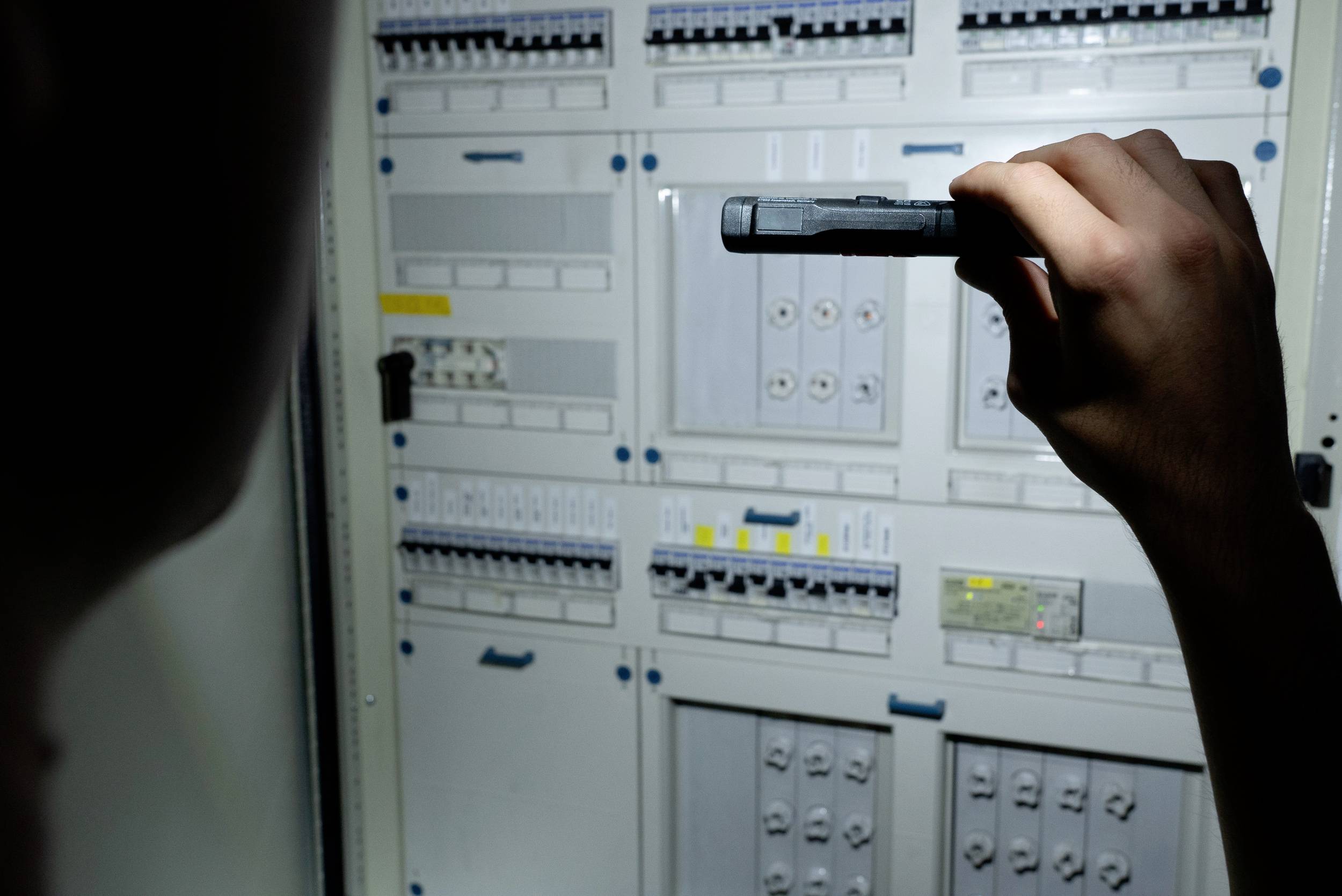 Osoba oświetla panel szafy sterowniczej latarką. Widocznych jest kilka przełączników i wskaźników.