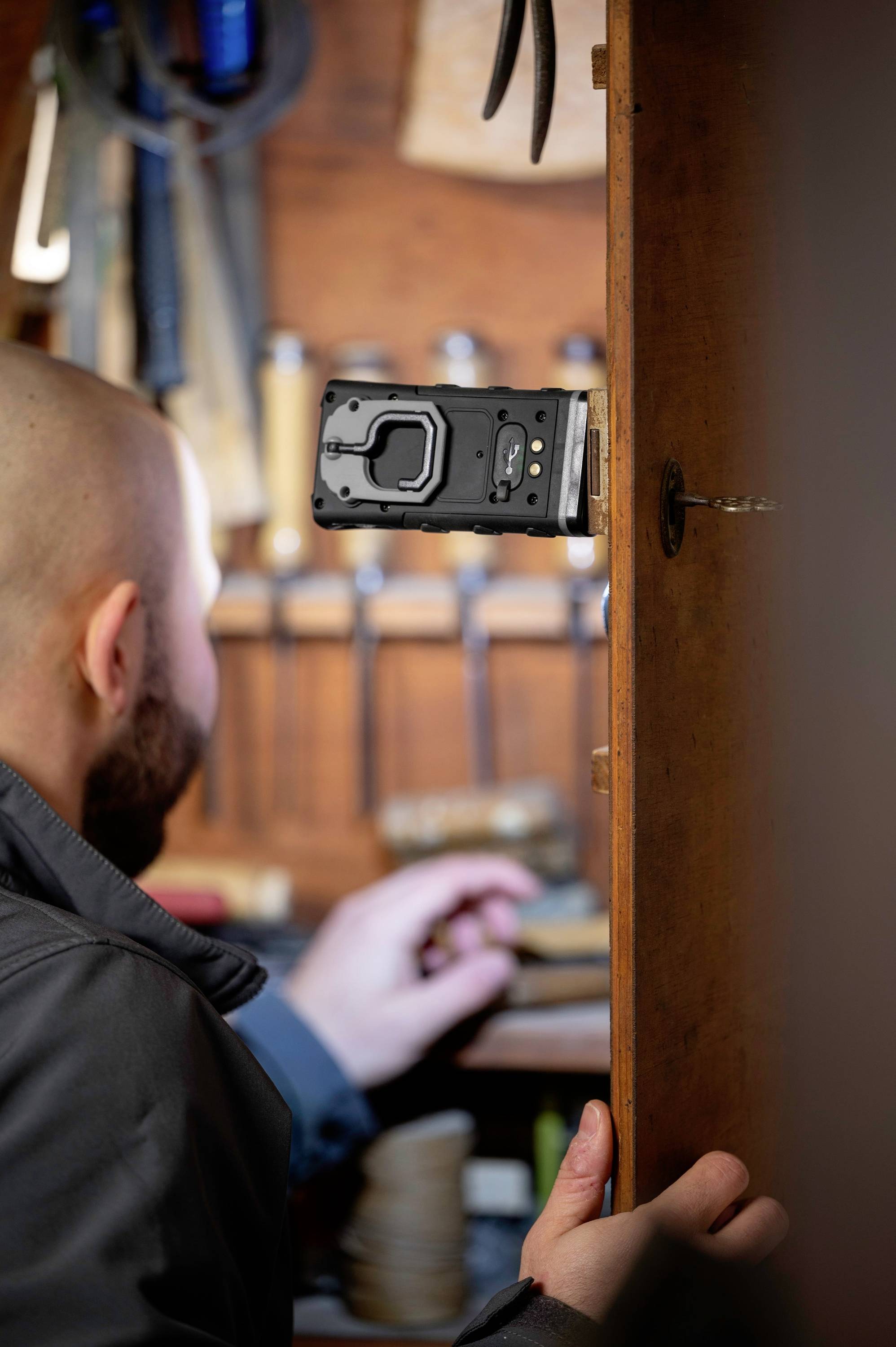 Mężczyzna pracuje w warsztacie stolarski. Czarne narzędzie jest przymocowane do drzwi, w tle widać inne narzędzia.