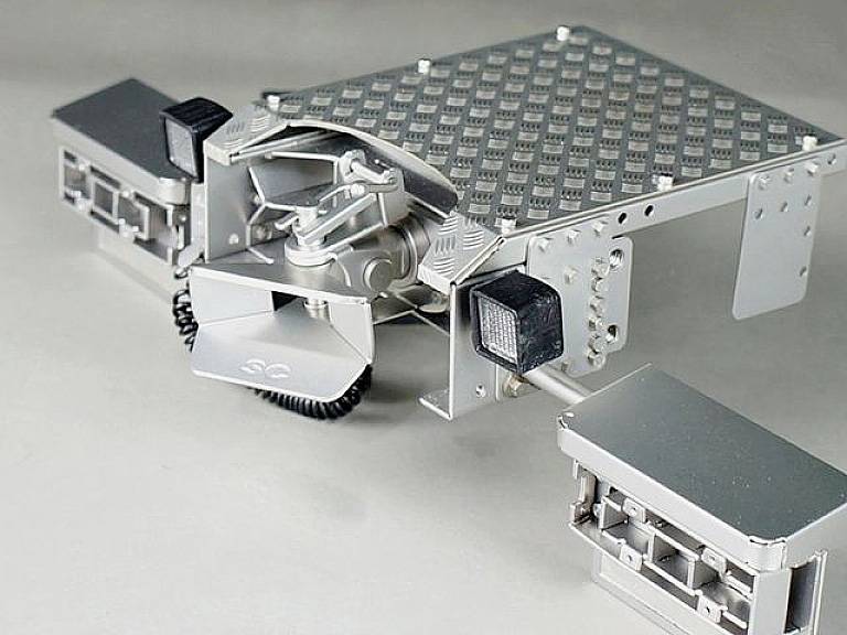 Srebrny, kwadratowy robot z czterema regulowanymi ramionami w futurystycznym warsztacie.