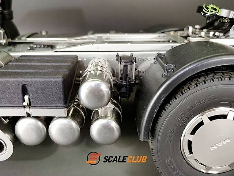 Zbliżenie modelu podwozia ciężarówki z metalowymi cylindrami i oponą. Logo 'ScaleClub' umieszczone na środku dolnej części obrazu.