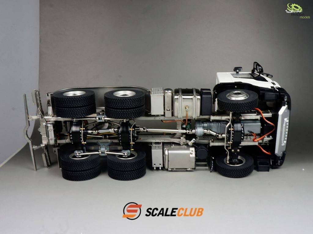 Miniaturowa ciężarówka widziana z boku przedstawia szczegółowe osie i koła od spodu. W prawym górnym rogu oraz na środku dolnej części widoczne są loga.