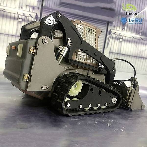 Mały zdalnie sterowany koparka gąsienicowy w hali. Jest szary z czarnymi elementami i posiada srebrną osłonę ochronną.