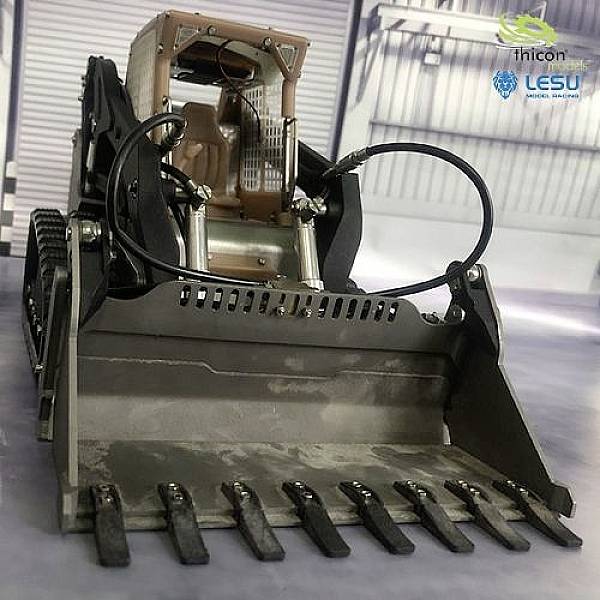 Mały koparka z łyżką i ramionami roboczymi stoi w warsztacie. Na tle widać różne maszyny i urządzenia.