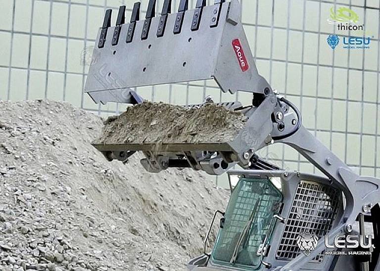 Koparka z łyżką podnosi ziemię przed ogrodzeniem siatkowym. Zdjęcie przedstawia zbliżenie mechanizmu łyżki.