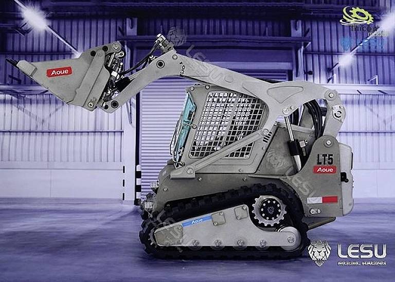 Kompaktowa, szara koparka z gumowymi gąsienicami podnosi łyżkę wewnątrz hali magazynowej. Widoczne są logo producentów.