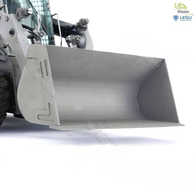 Szary ładowarka kołowa z szeroką łyżką jest widoczny od przodu. Stoi na jasnej powierzchni. W tle widoczne są niektóre komponenty maszyny.